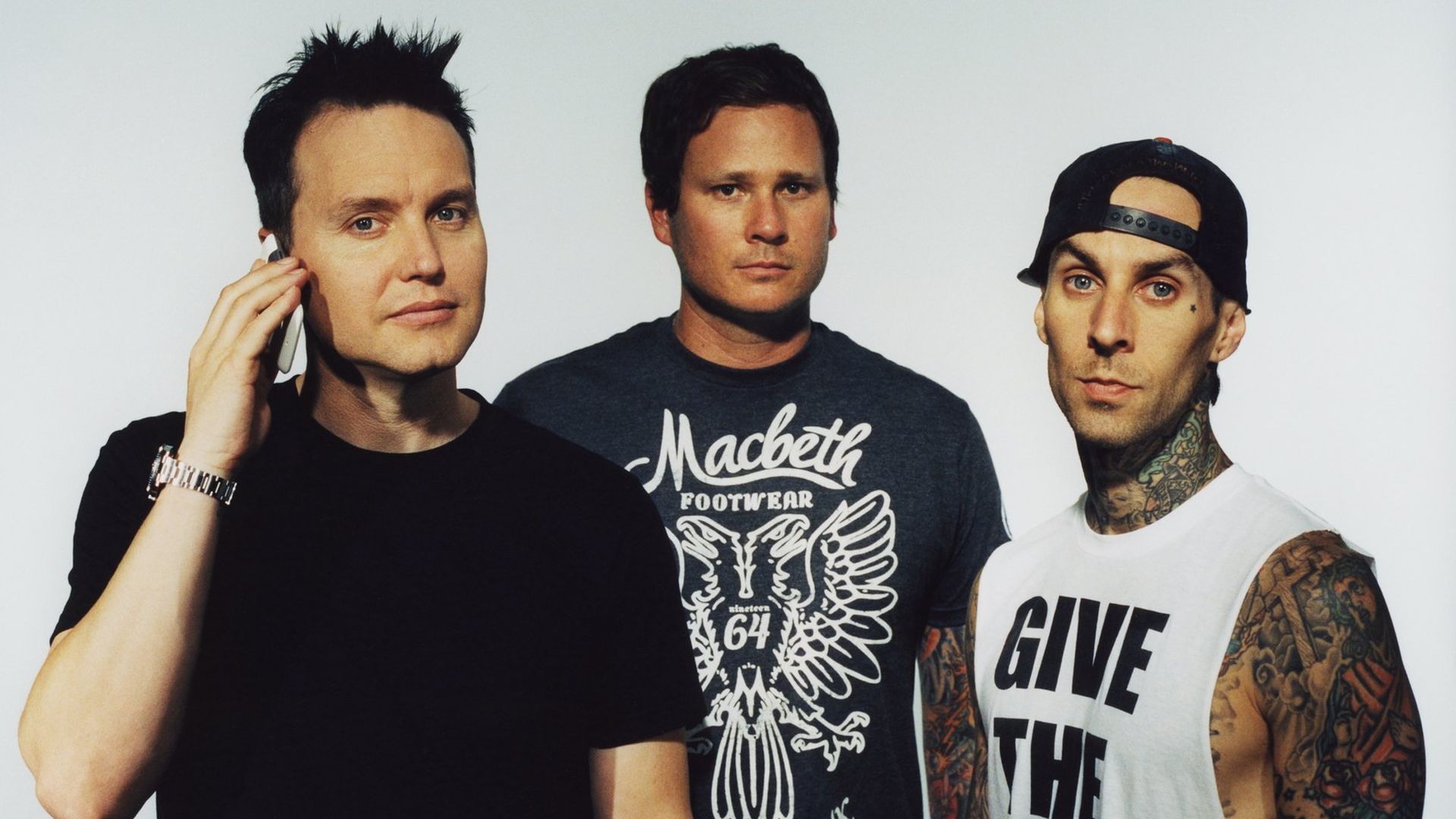 Three men pose in a polaroid-style frame against a white background: left man on a phone, center wearing a dark tee with the "Macbeth" crest, right tattooed in a white tank top with bold text.