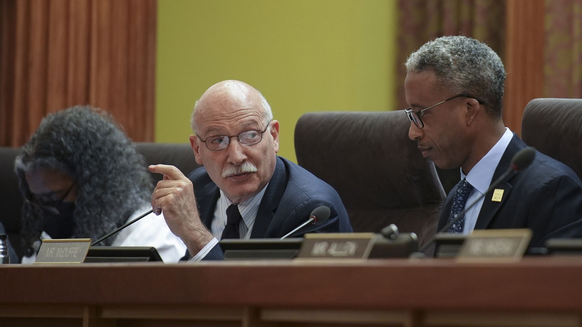 Three officials seated at a panel desk in a meeting room; a Phil Mendelson speaks while gesturing
