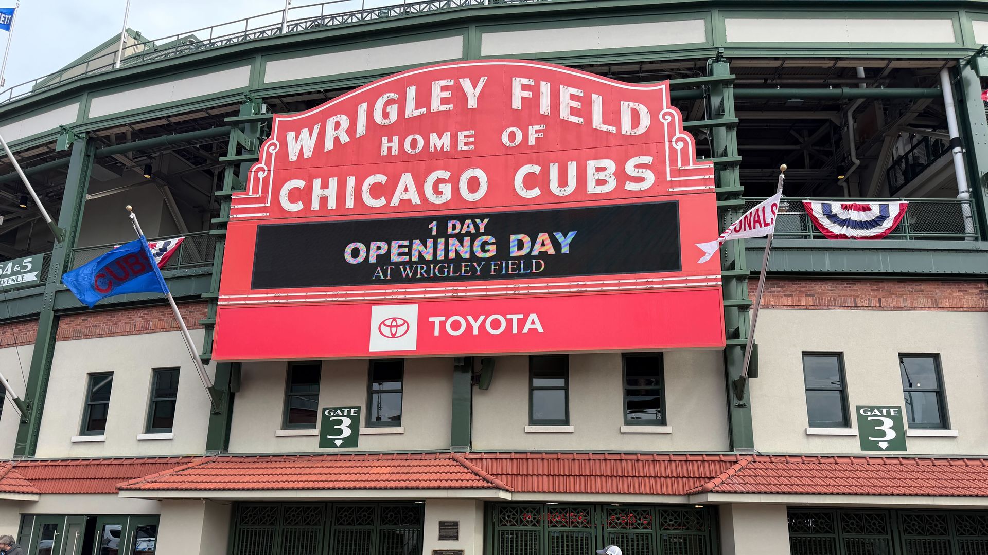 Wrigley Field exterior with a red sign, a digital marquee reading '1 Day Opening Day at Wrigley Field', and a Toyota banner, as a person in white top and maroon pants walks on brick by green gates.