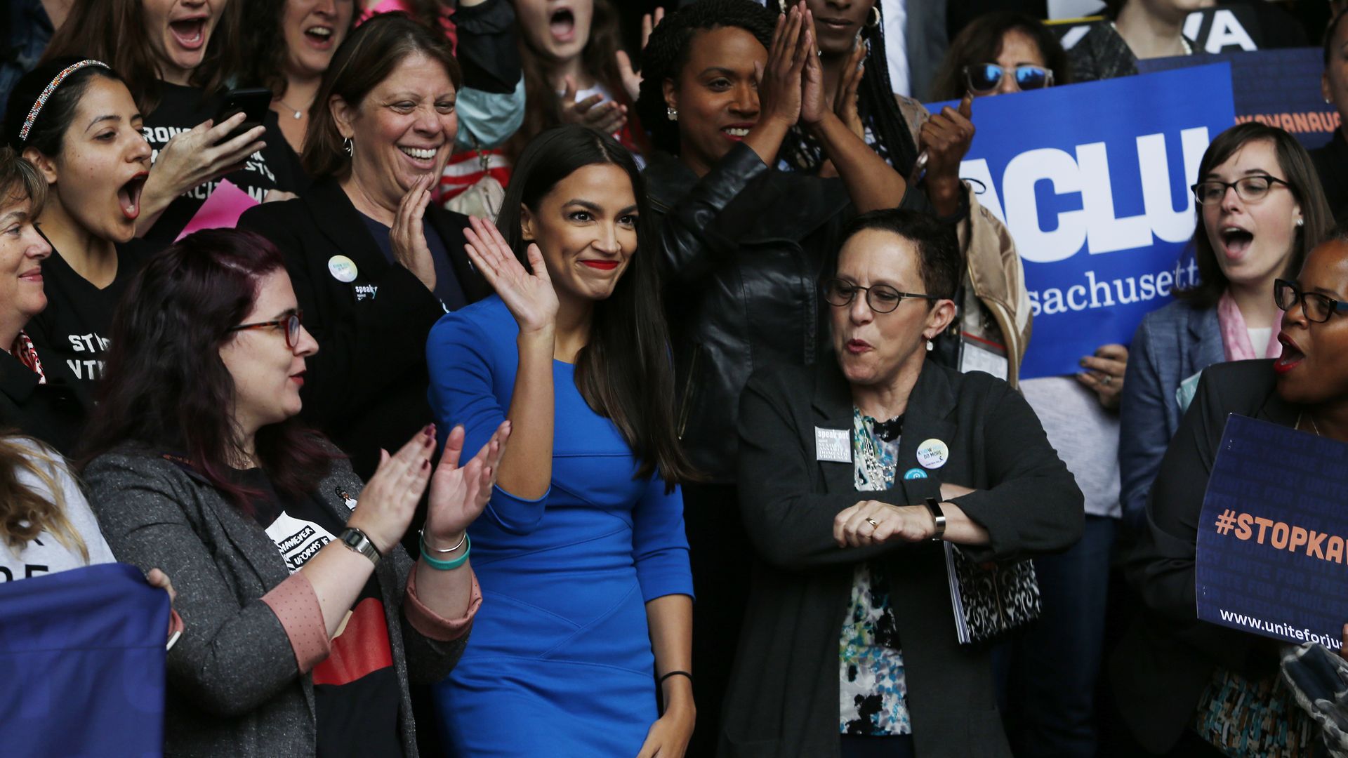 Alexandria Ocasio-Cortez waves in the midst of a crowd wearing a bright blue dress.