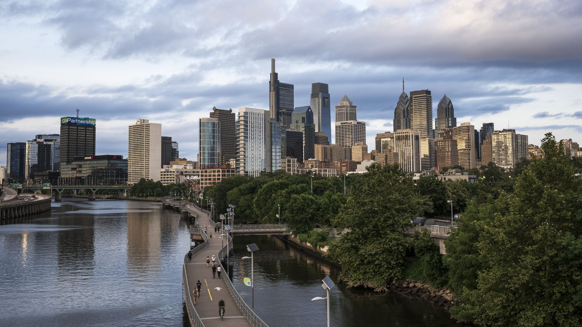 City skyline with tall modern skyscrapers along a wide river. A curved pedestrian promenade runs beside the water, with a bridge on the left and lush trees lining the shore under a cloudy blue sky.