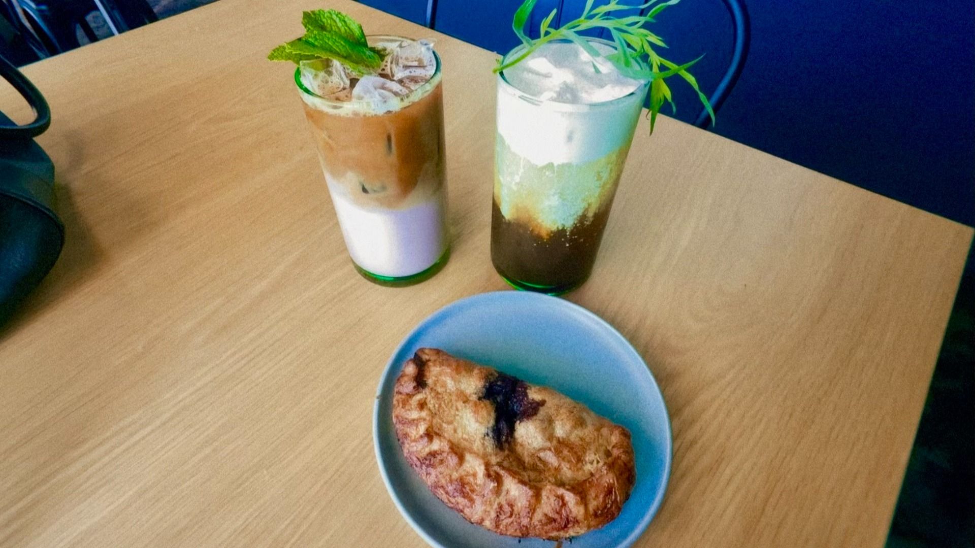 Two tall, layered iced drinks with mint garnish on a light wood table; a blue bowl holds a slice of pie in the foreground, and blue chairs are in the background.