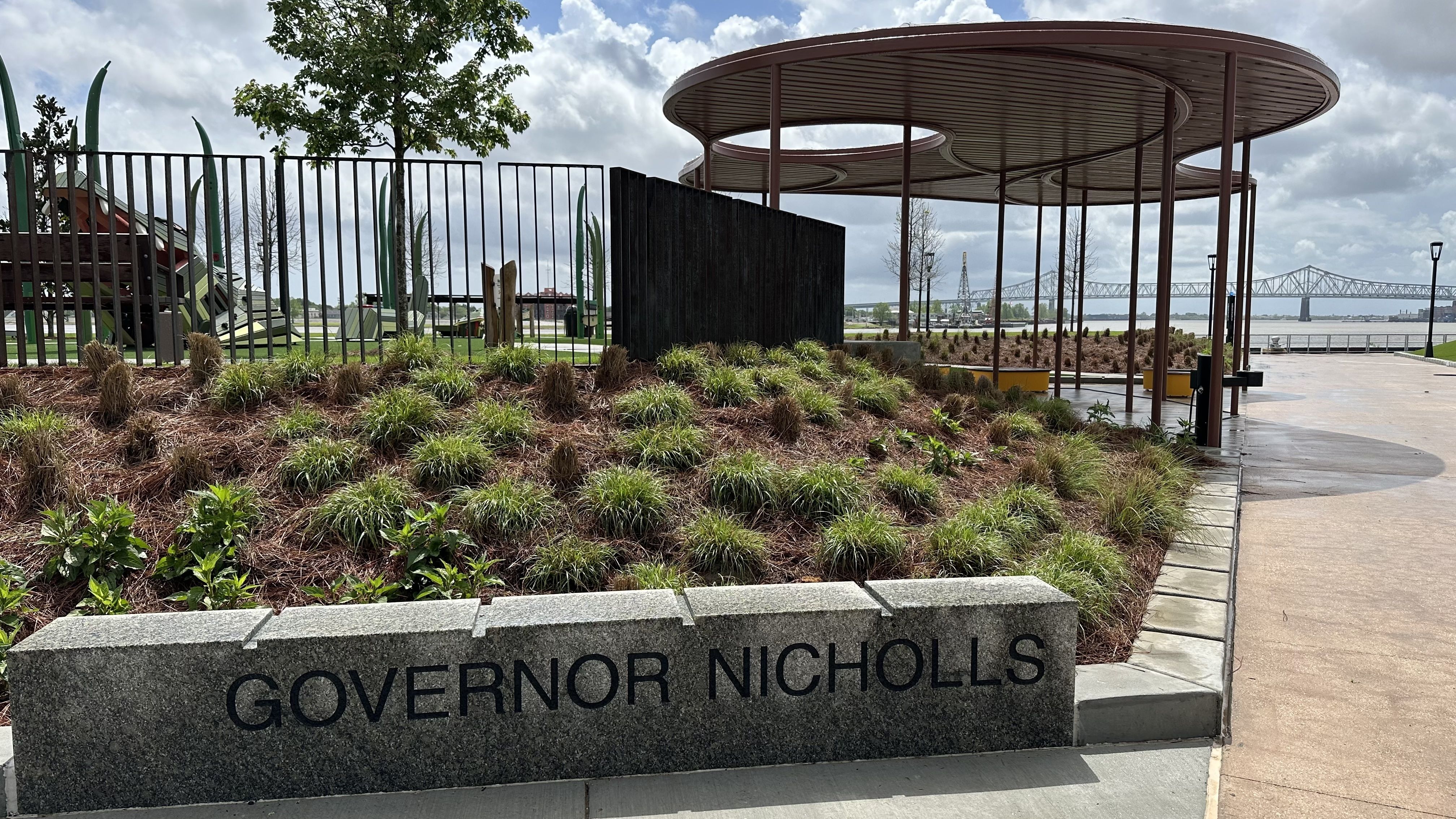 Park by the river with a stone sign reading "GOVERNOR NICHOLLS", a black metal fence, manicured shrubs, a circular shaded pavilion, a paved walkway, and a distant bridge under a cloudy sky.