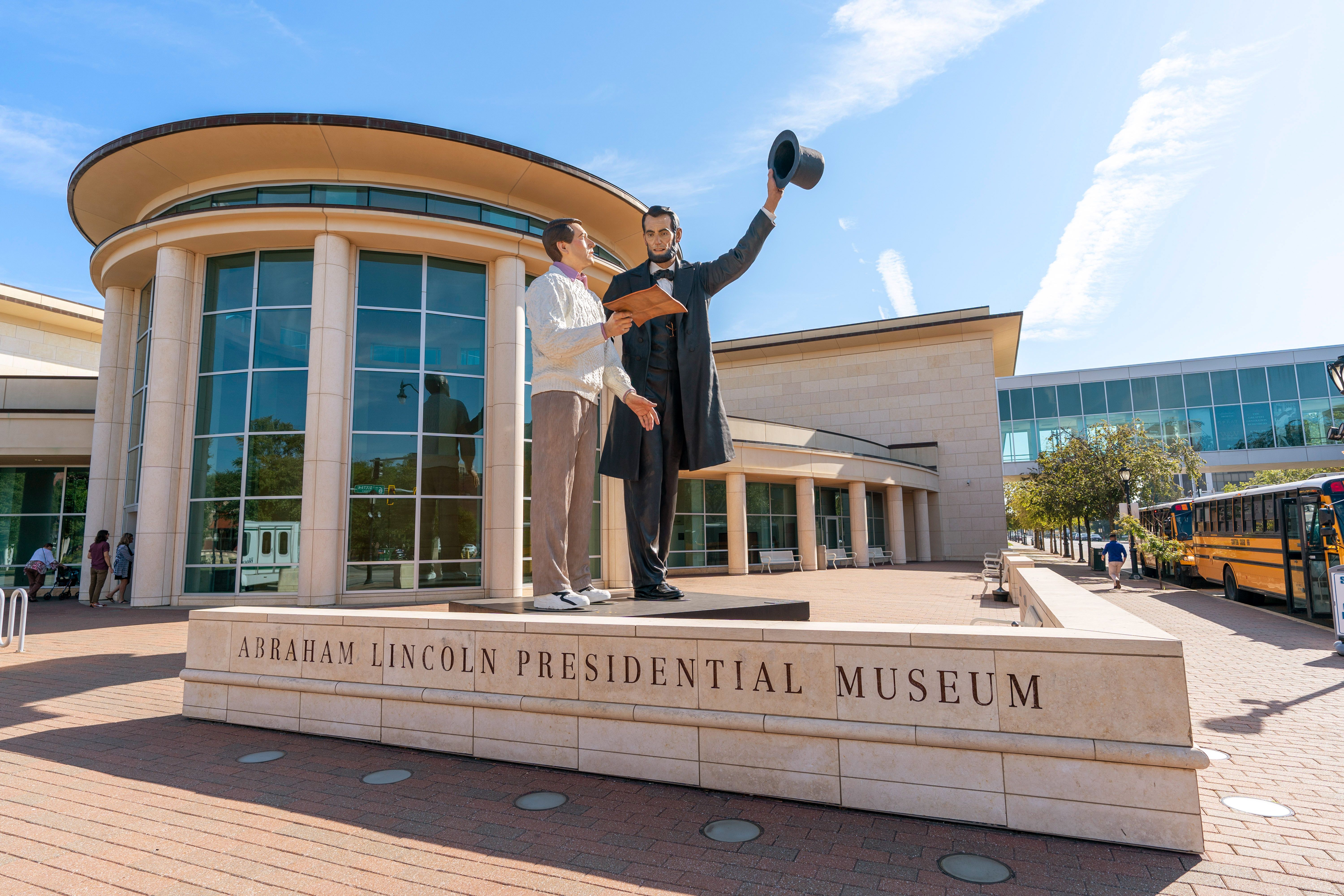 Two statues outside the Abraham Lincoln Presidential Museum: a dark-coated figure raises a top hat while a lighter-clothed figure holds a folder; glass facade, clear blue sky, and a yellow school bus nearby.