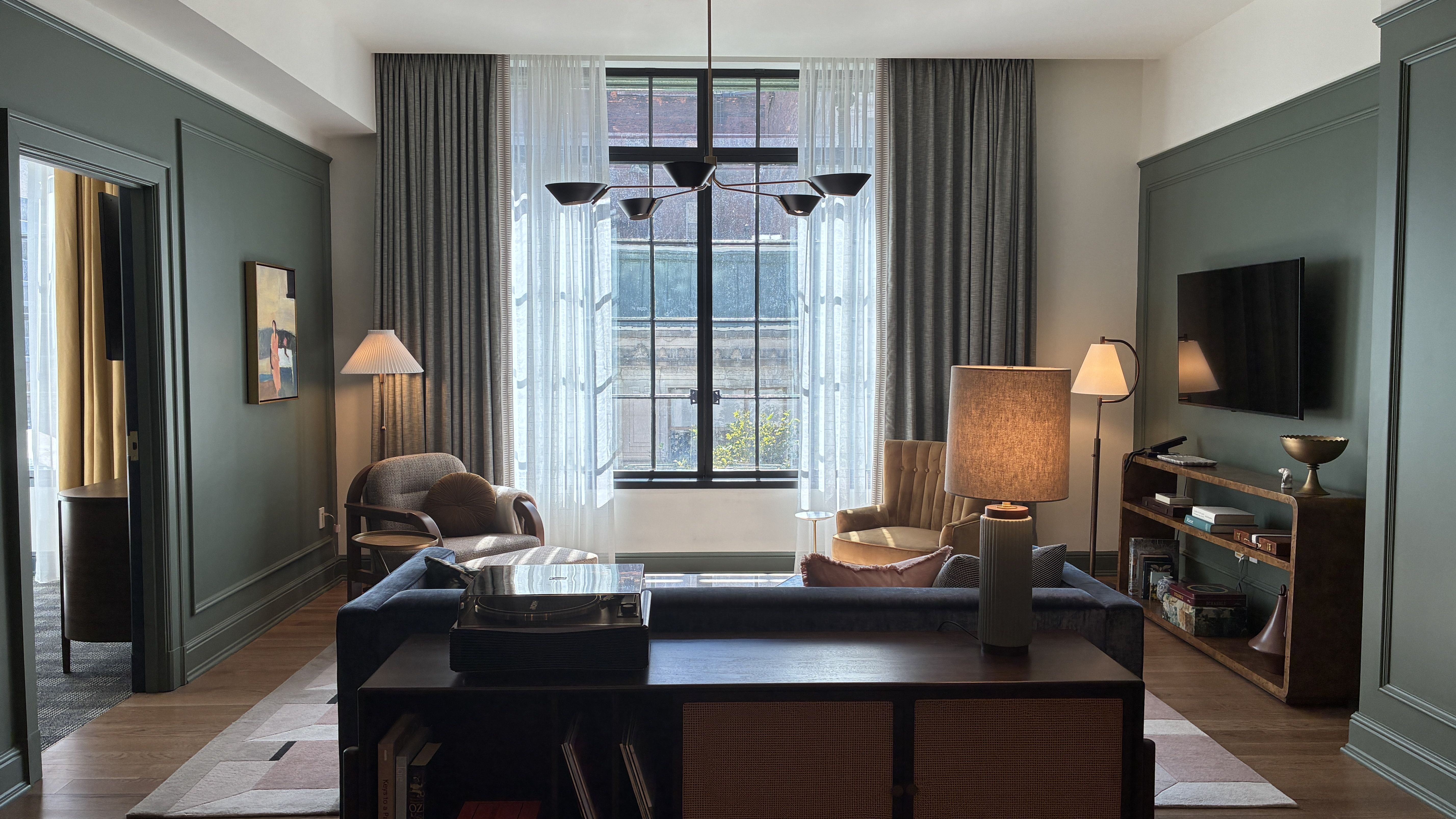 Teal-walled living room with a large window, sheer gray curtains, a black multi-arm chandelier, brown seating, a coffee table with a turntable, a wall-mounted TV, and a bookshelf.