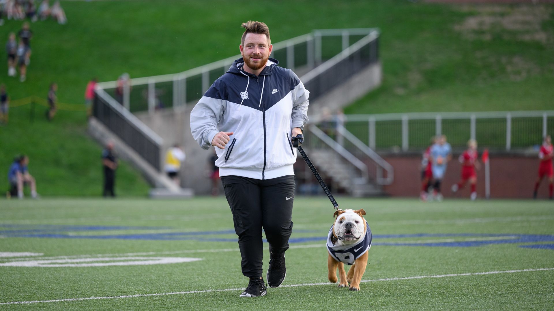 A man in a black and white Nike jacket walks on a green sports field holding a leash attached to a bulldog wearing a matching Nike sweater. Spectators and players are in the background.
