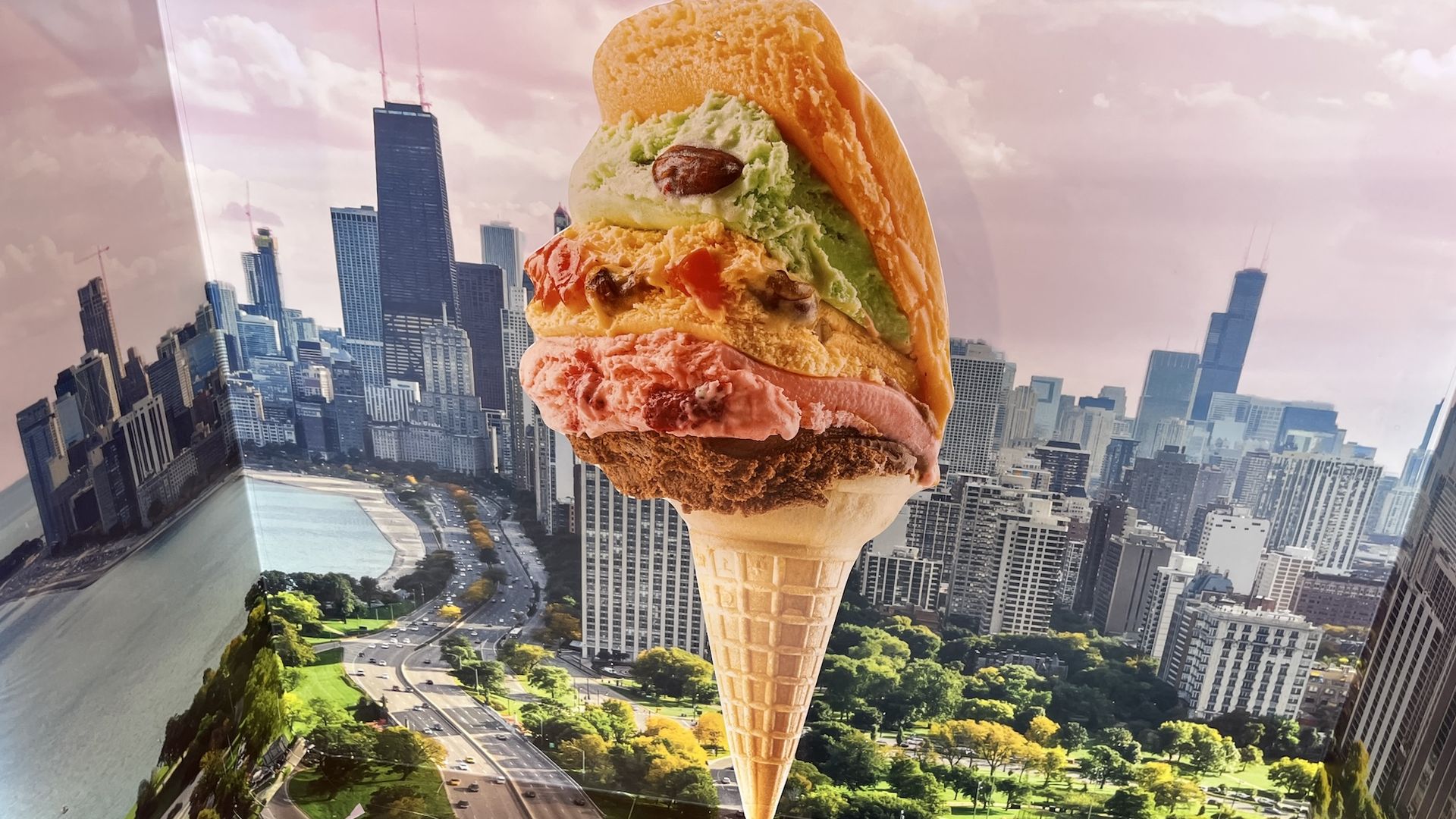A large multi-scoop ice cream cone with pink, green, orange, and vanilla scoops stacked on a waffle cone, set against a pink-tinted city skyline and highways.