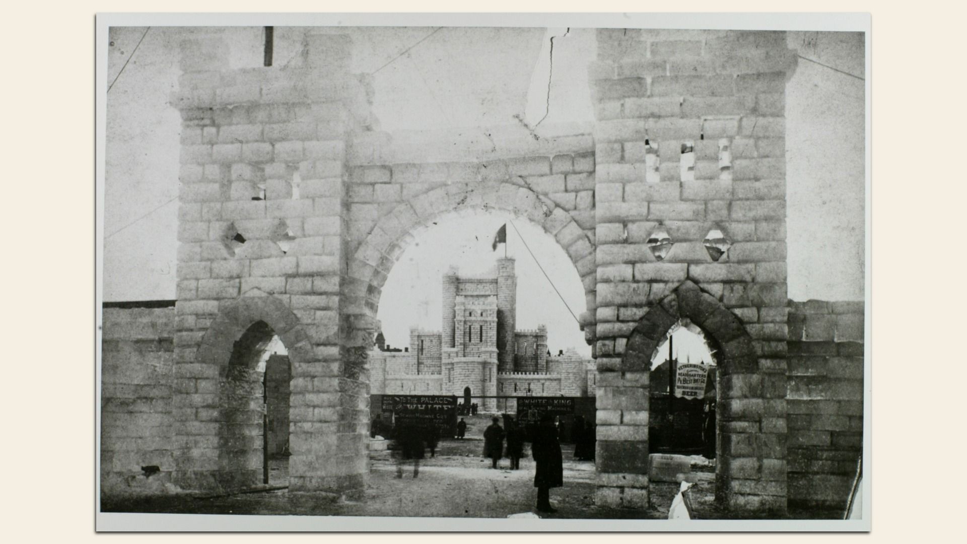 Black and white photo of a stone castle gate with two towers and an arched entrance, people walking through, and a larger castle structure visible in the background.