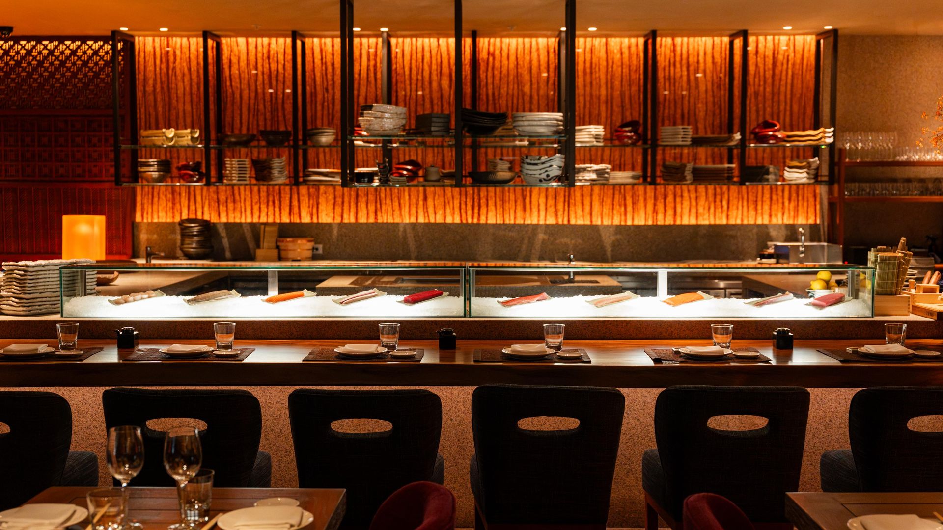 Warm, orange-lit sushi bar with a glass display of nigiri on ice, shelves of plates behind wooden panels, and tables set with dishes in the foreground.