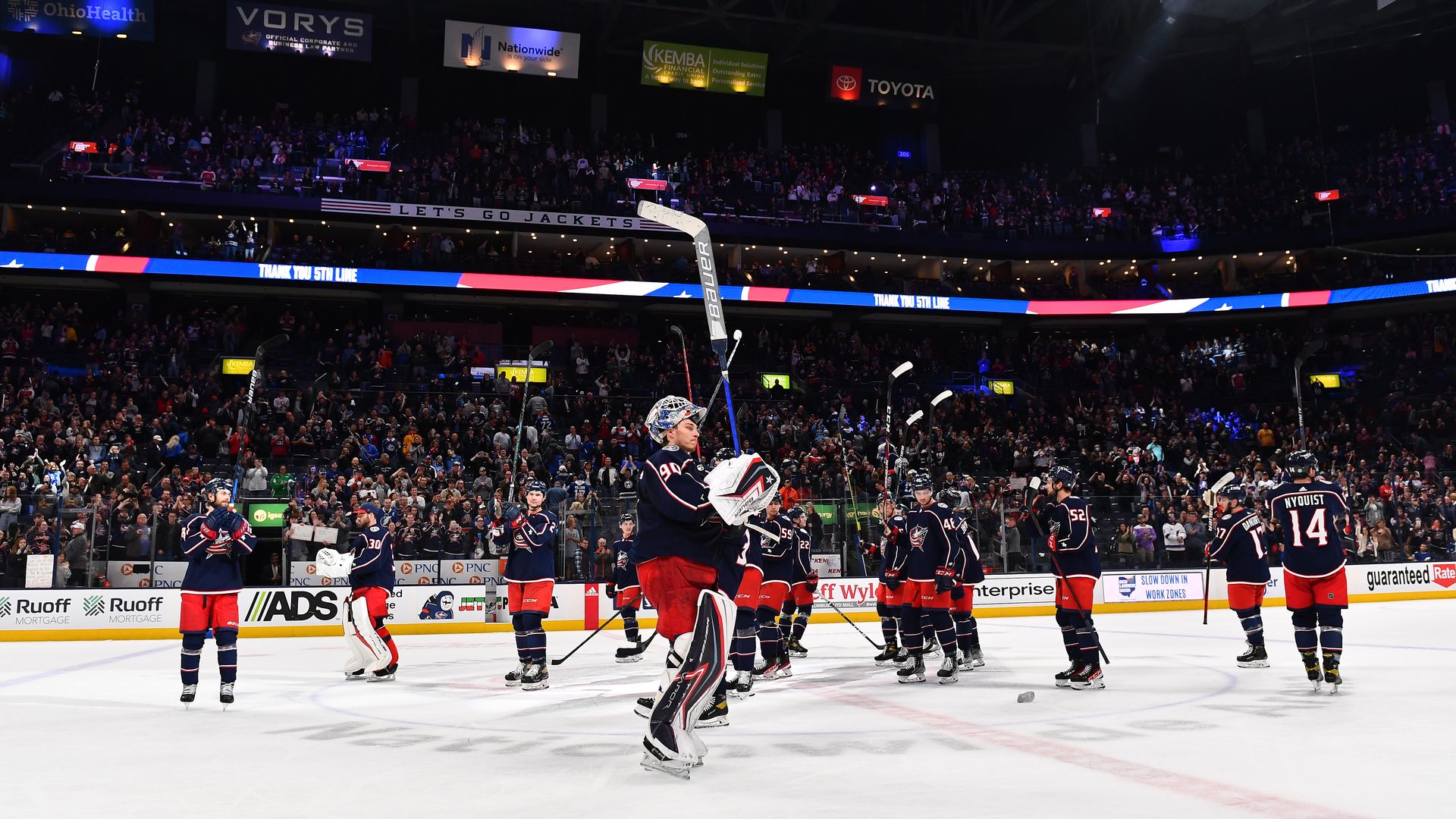 Columbus Blue Jackets raising their hockey sticks and saluting fans from the ice at Nationwide Arena.