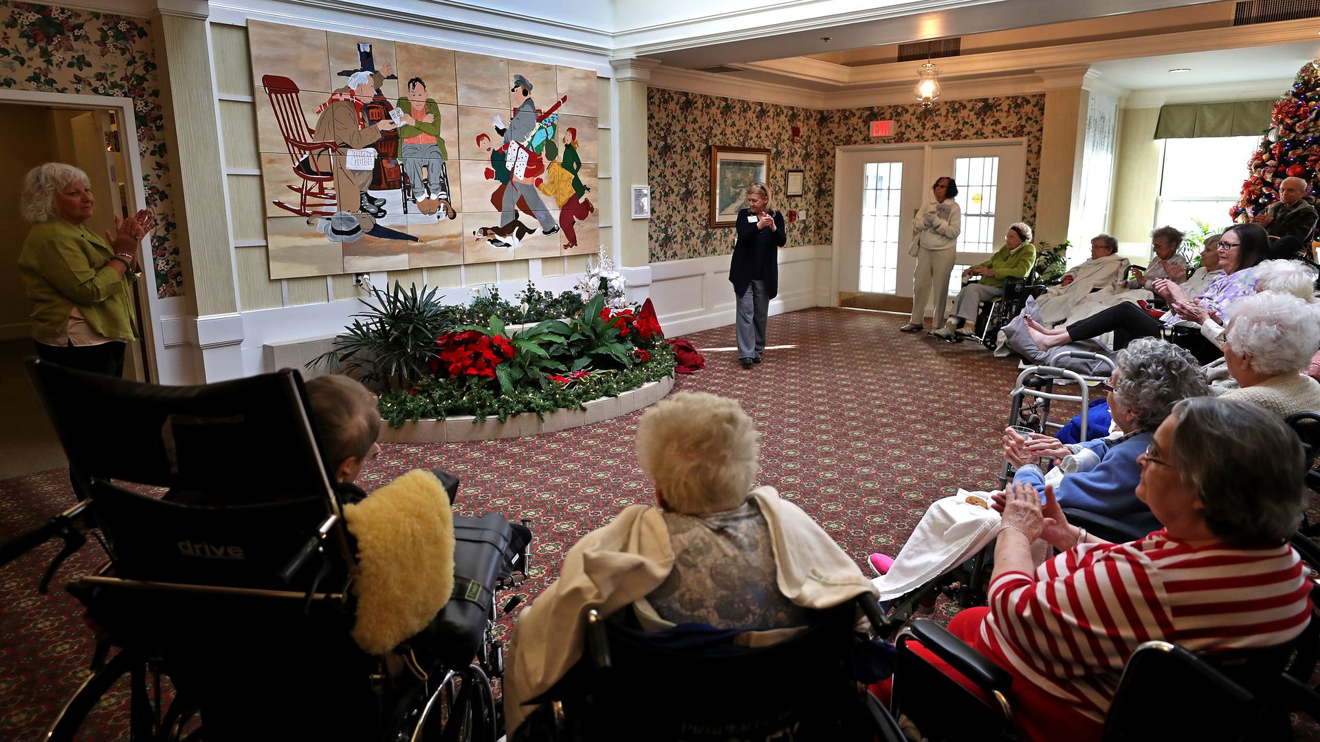 A group of elderly people sit in a nursing home commons area.