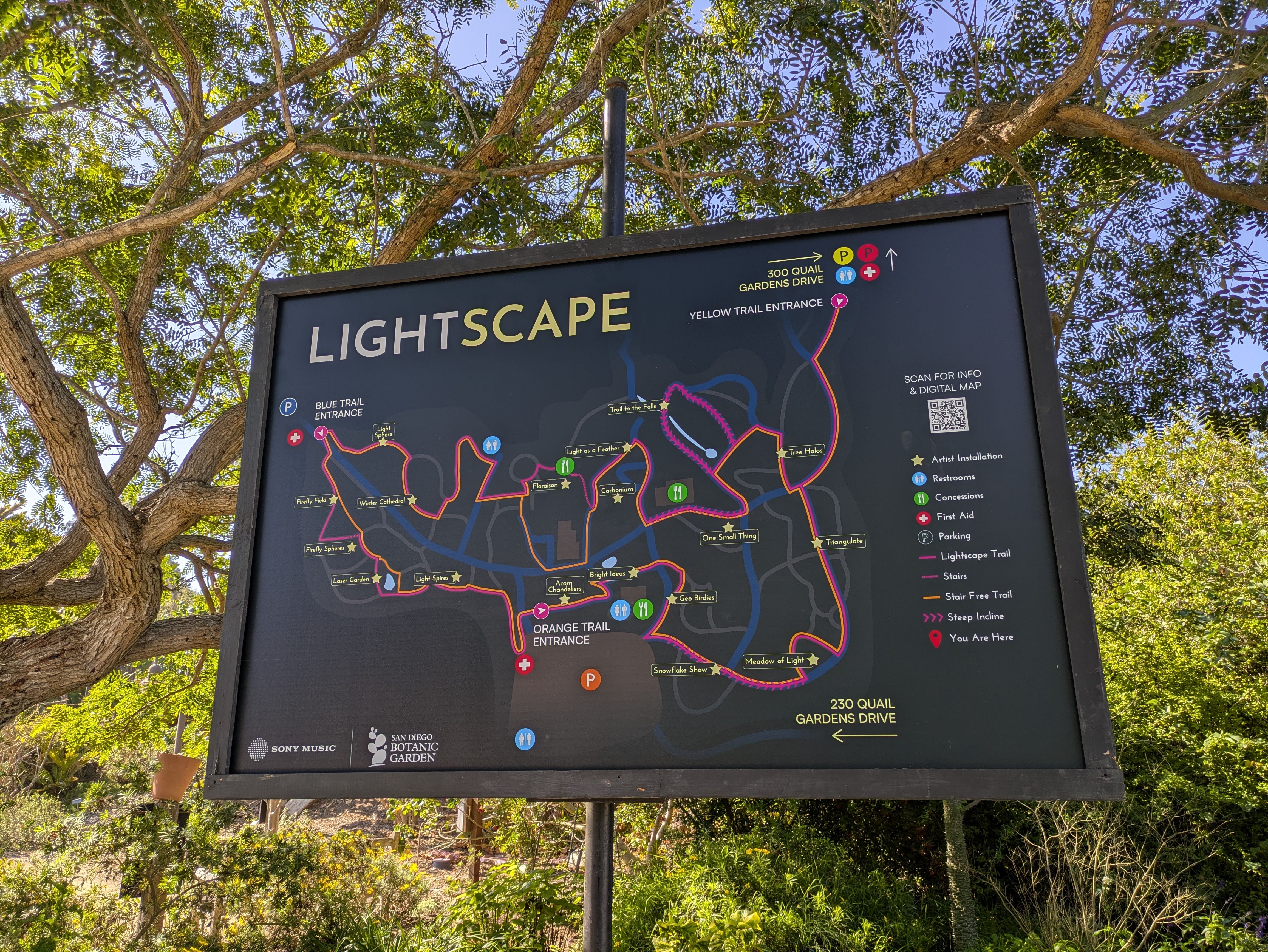 Map of Lightscape trails at San Diego Botanic Garden with blue, yellow, and orange trail entrances, artist installations, restrooms, concessions, first aid, and parking marked.