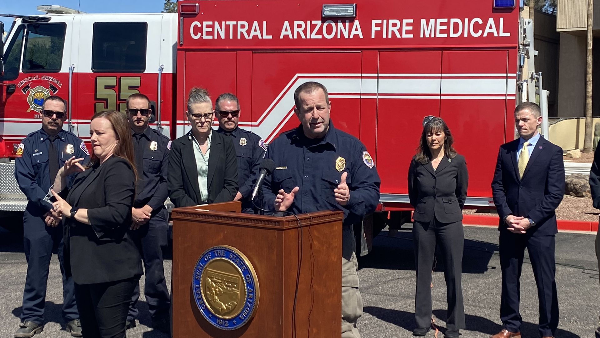 A man in a blue uniform and a badge gestures from behind a podium while standing in front of a fire truck.