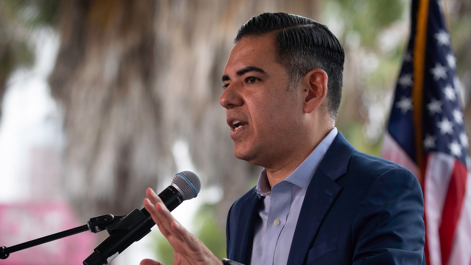Rep. Robert Garcia, wearing a blue suit and speaking into a microphone in front of an American flag outdoors.