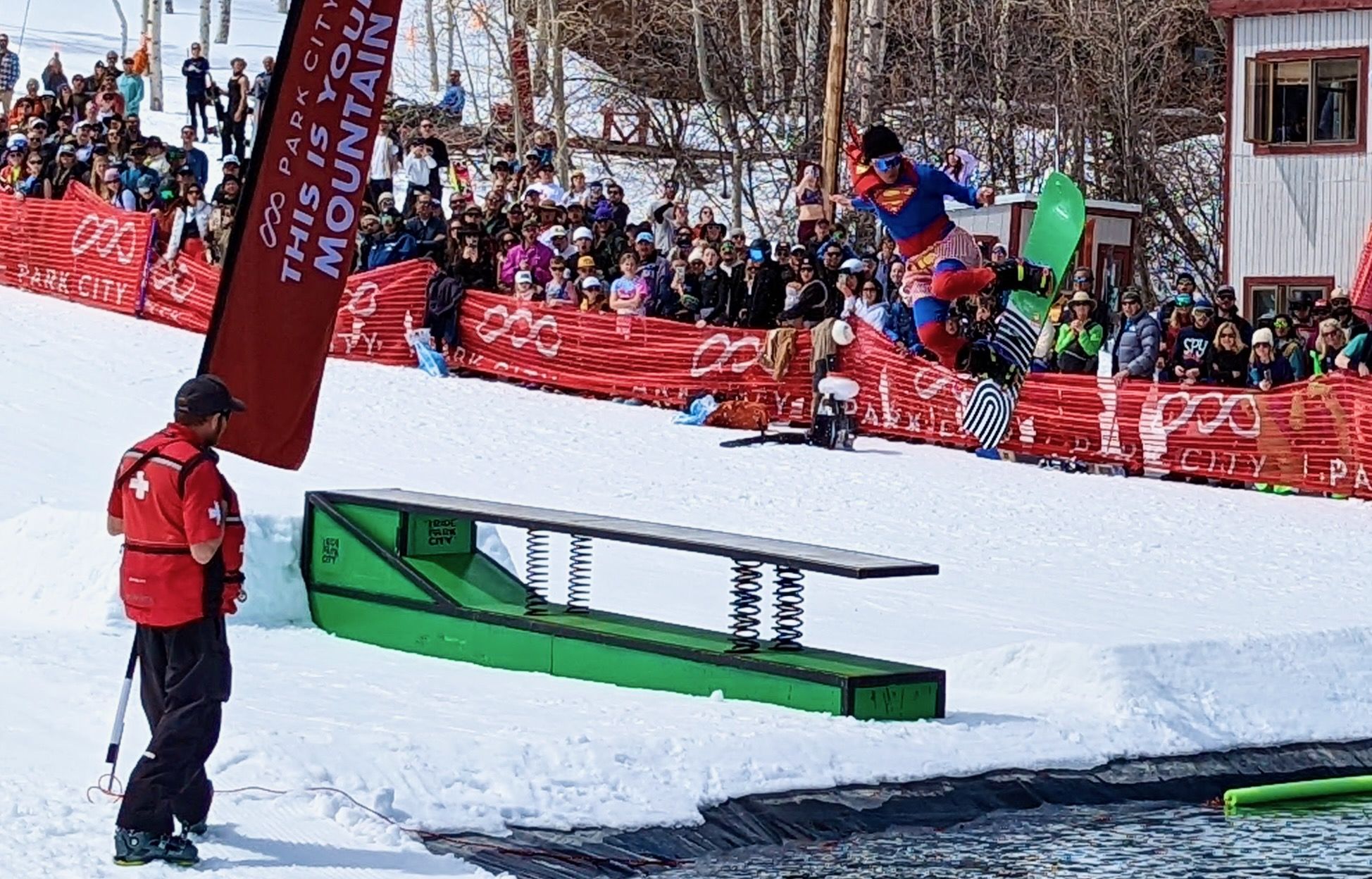 A skier dressed as Superwoman vaults off a ski jump over a pool of water.