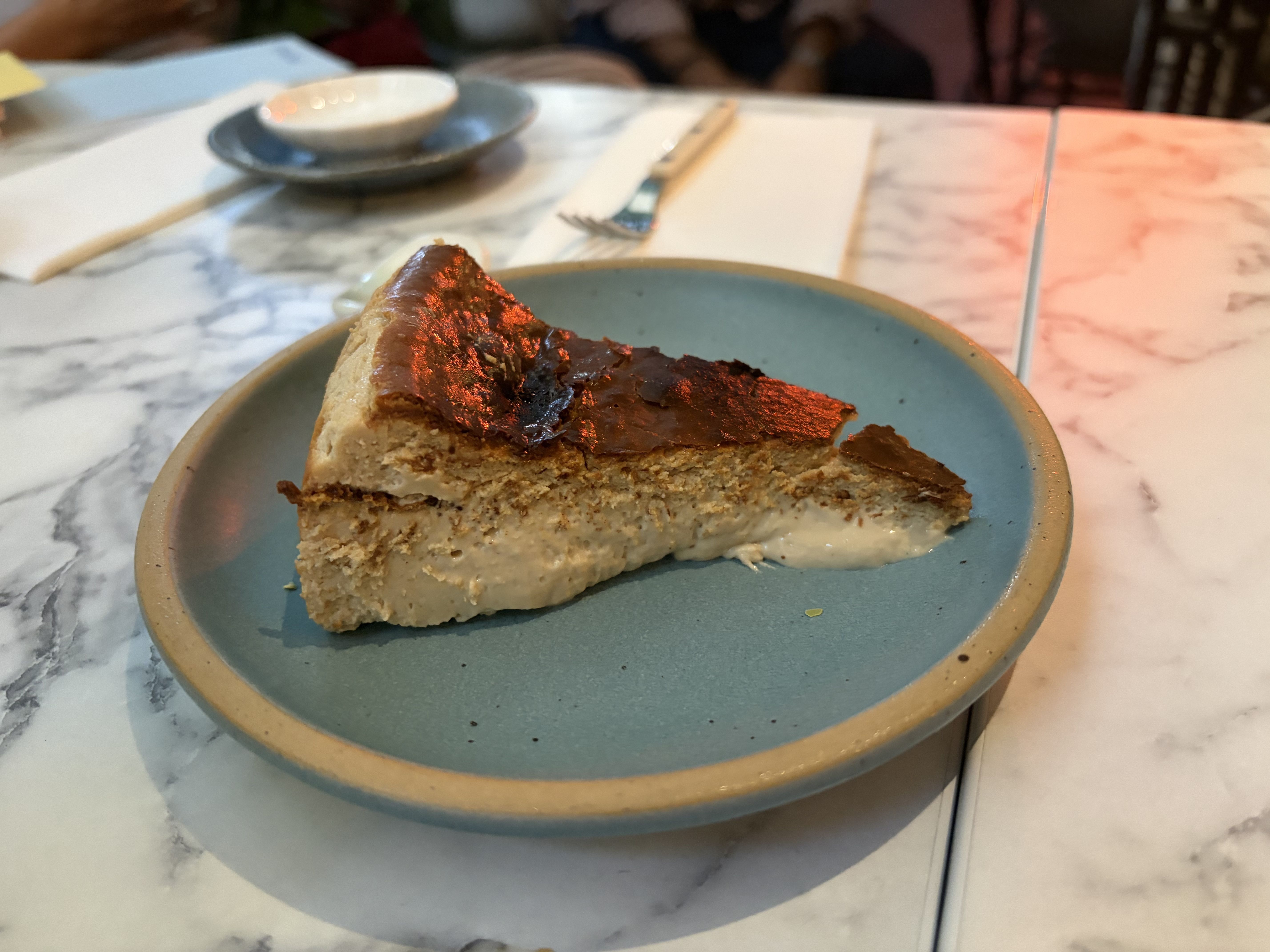 A slice of cake with a glossy chocolate topping on a blue plate rests on a marble table. 