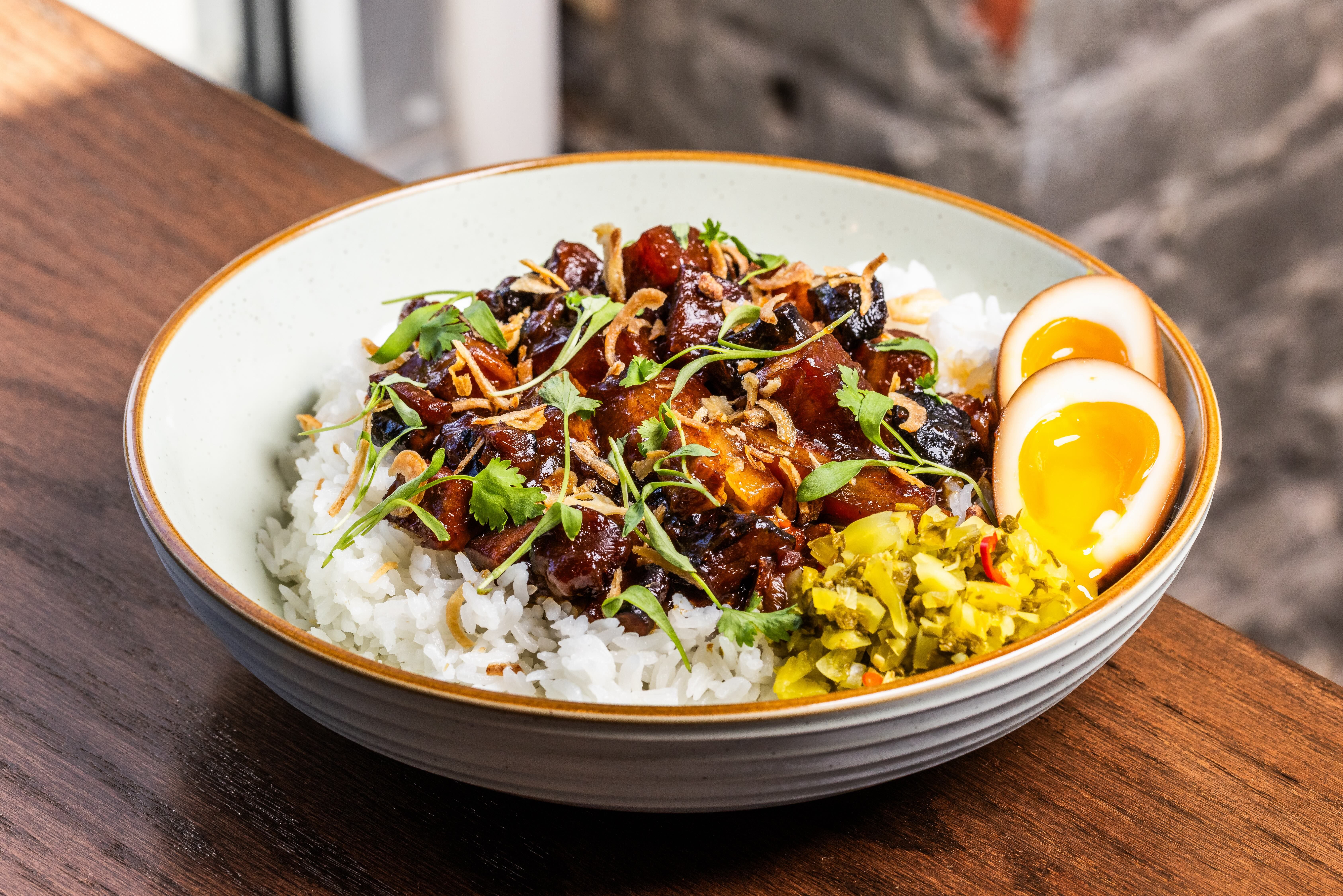 Bowl of white rice topped with glossy meat, crispy onions, cilantro, and a halved soft-boiled egg; pickled greens on the side, all set on a wooden table.