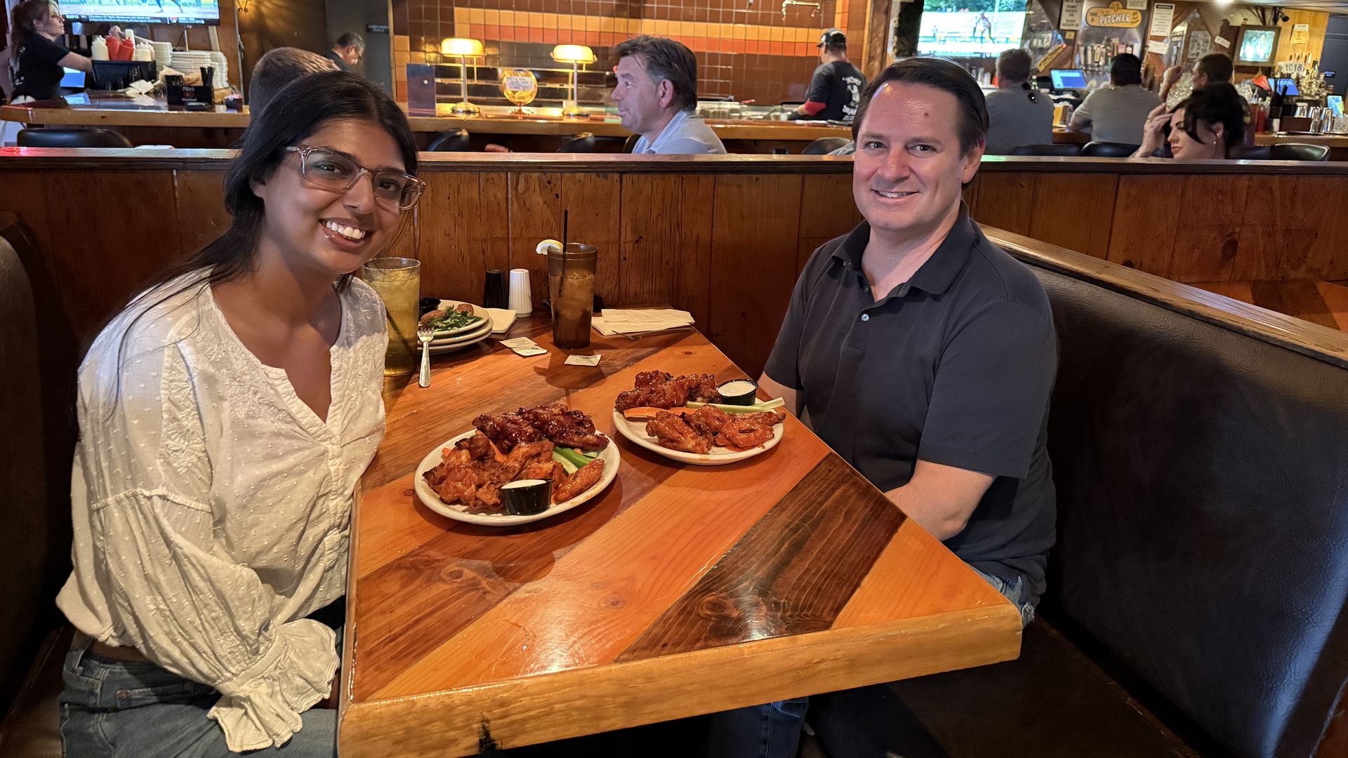 Two friends sit at a wooden booth in a busy sports bar, smiling at the camera. Plates of wings and drinks on the table, with TVs showing sports and a colorful round sign behind the bar.