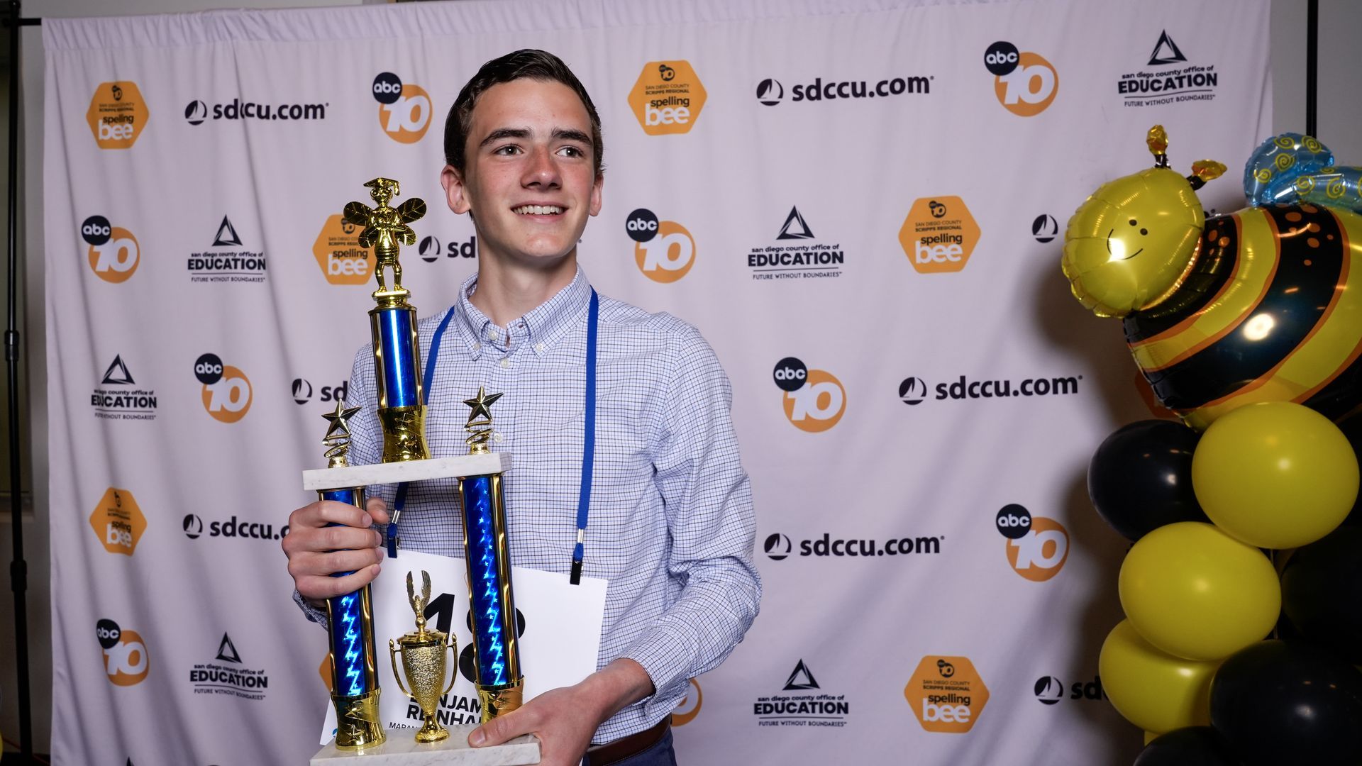 A teenage boy in a blue button-down shirt smiles while holding a trophy with blue columns and gold tops, in front of a sponsor backdrop with yellow bee balloons on the right.