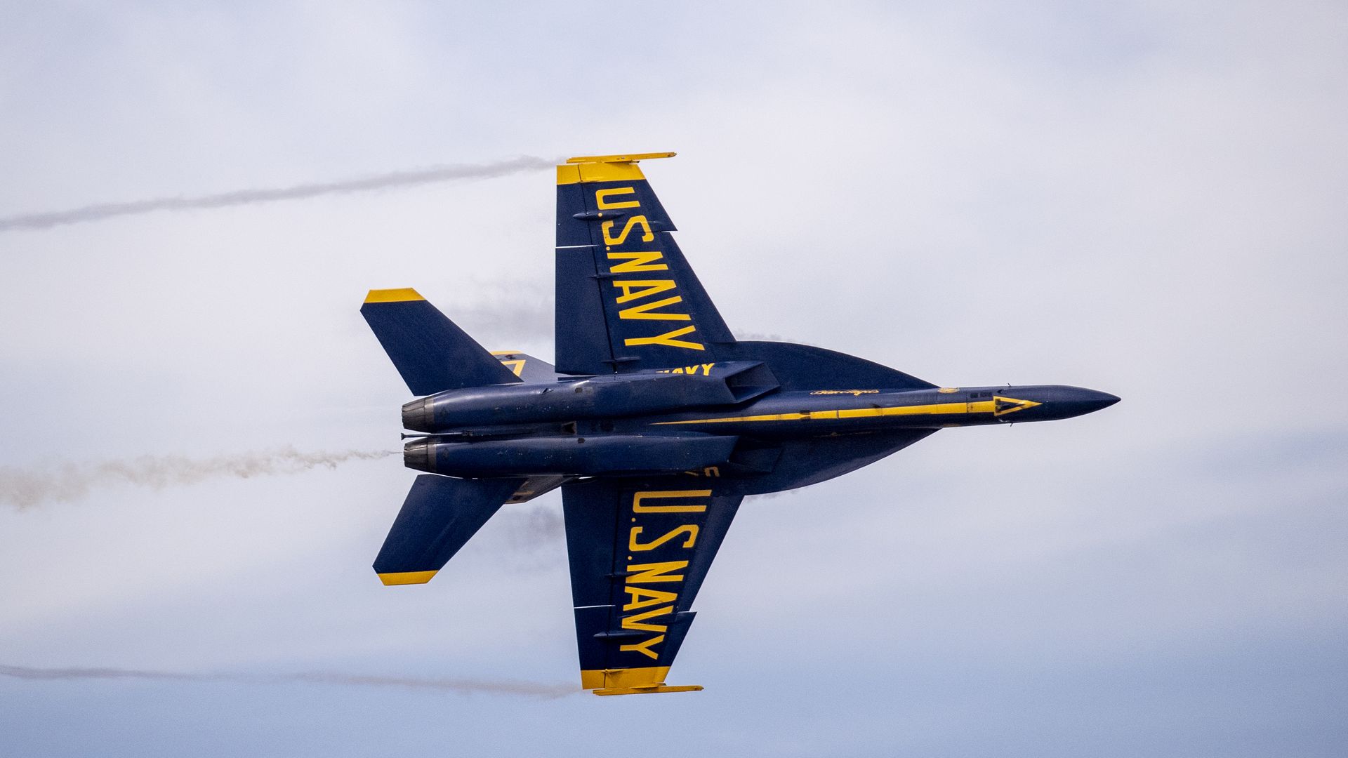 Blue and yellow US Navy fighter jet flying with afterburners on, leaving faint smoke trails against a cloudy sky.