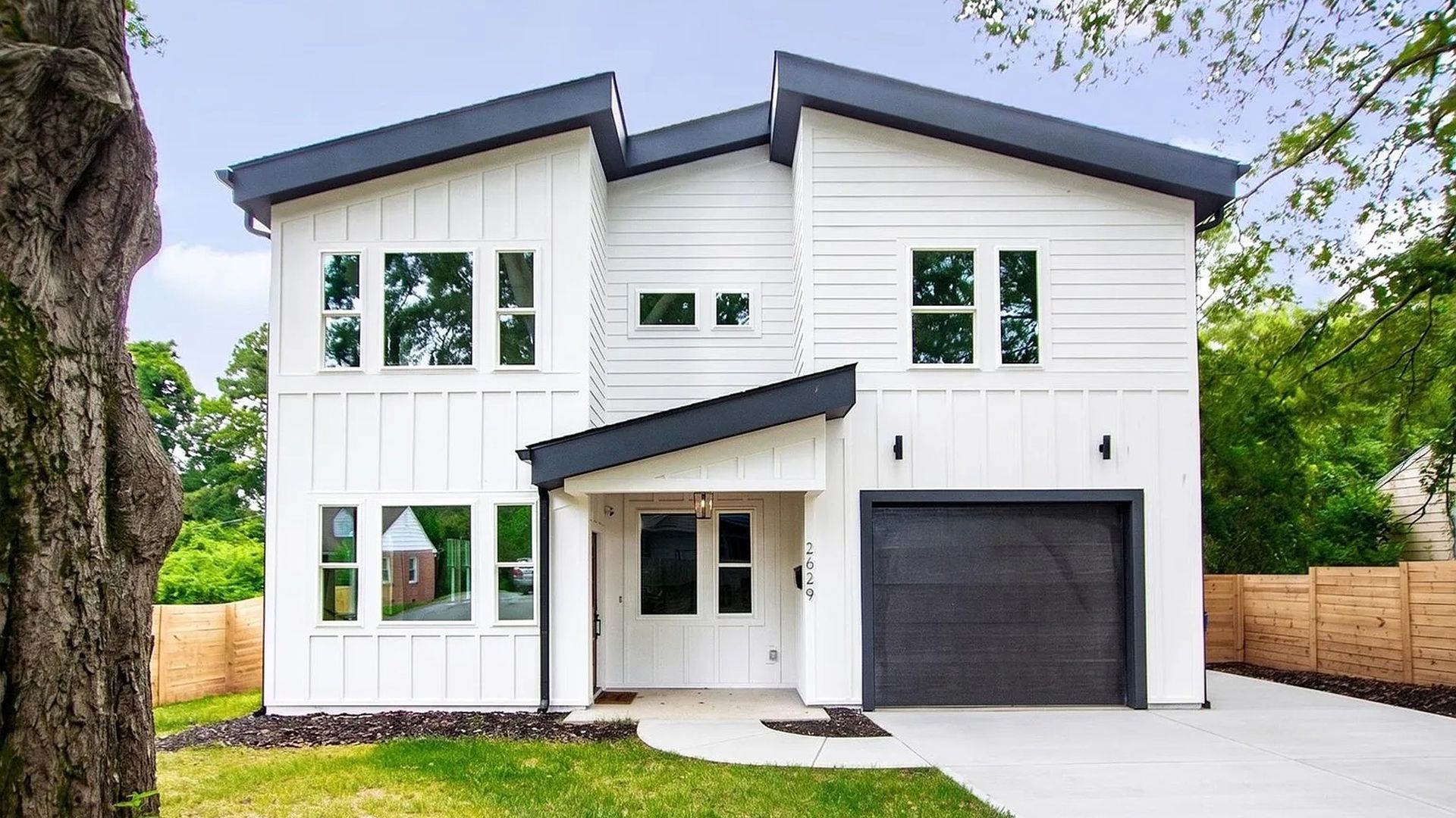 Modern two-story white house with dark gray roof edges, large first-floor windows, a small covered entry, and a gray garage door. Green lawn and trees frame the front yard with a wooden fence on the side.