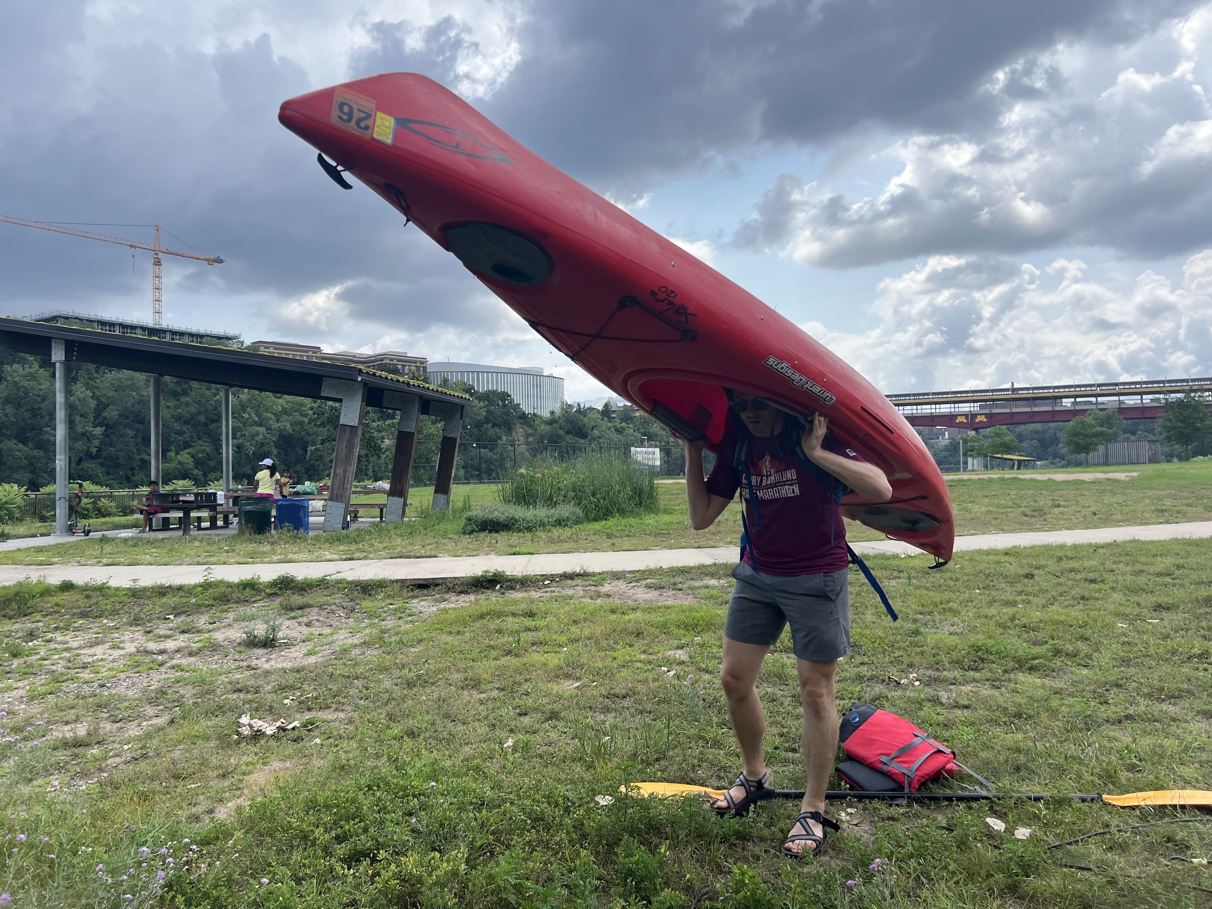 The dweebish looking man from an earlier image putting a kayak on his shoulders — which is not how you carry a kayak — on a grassy field with a paddle and life jacket at his feet.