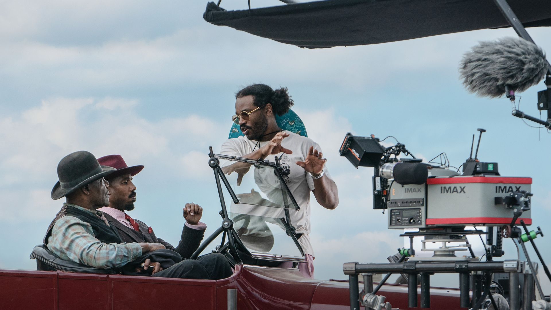 Two men in vintage clothing sitting in a red open-top vehicle while a man with sunglasses and a white t-shirt directs beside an IMAX camera under a cloudy sky.