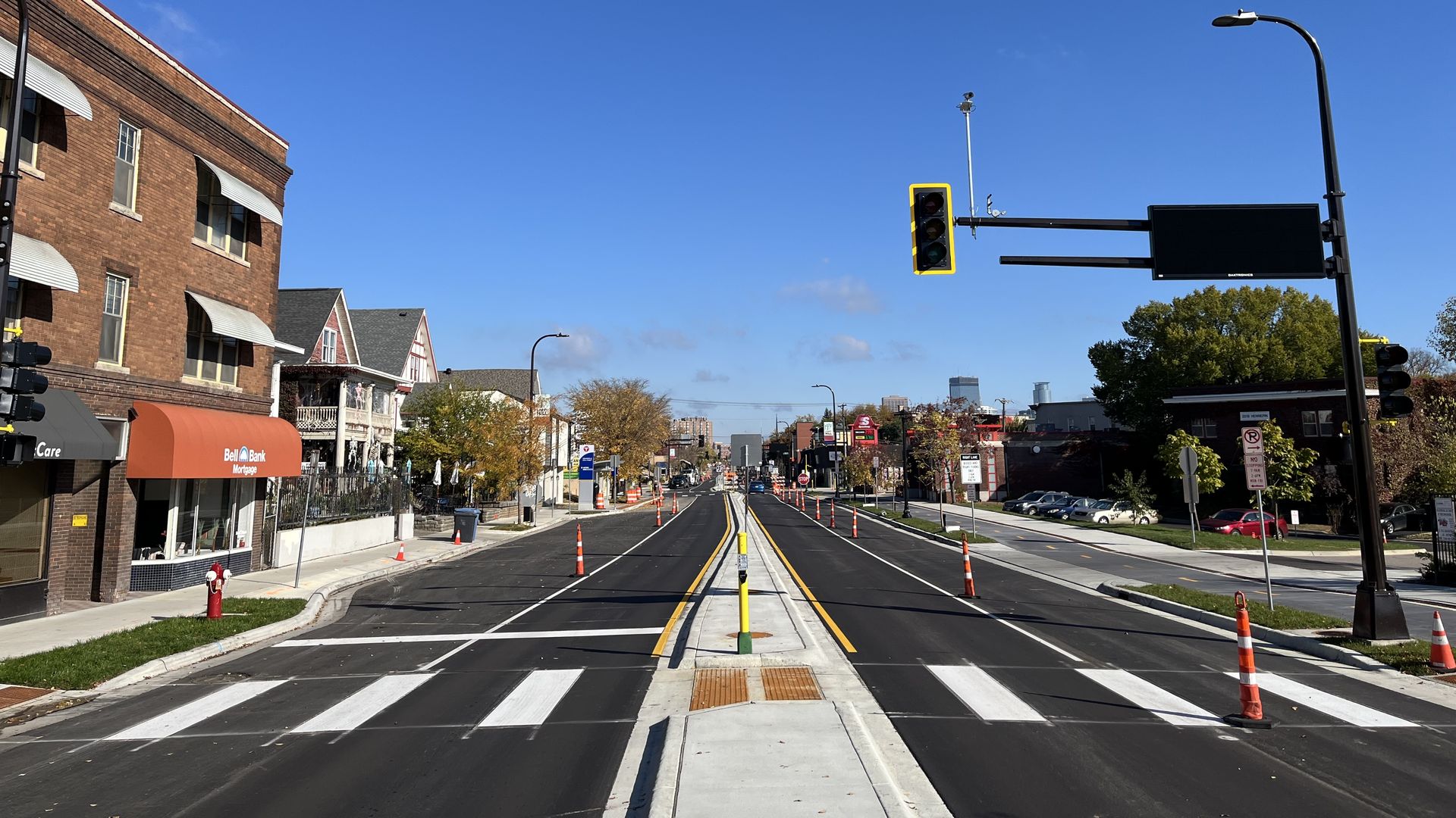 Empty newly paved city street with orange traffic cones, crosswalks, traffic lights, brick buildings, storefront with orange awning, and clear blue sky on a sunny day.