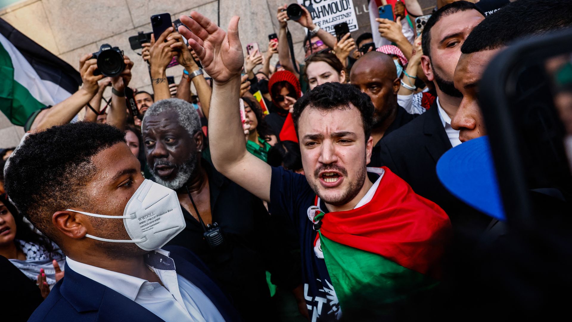 A black-haired, trim-bearded former Columbia University student Mahmoud Khalil, wearing a black t-shirt with a white symbol and a red and green flag tied around his neck, holds his  right hand aloft while speaking in a crowd.