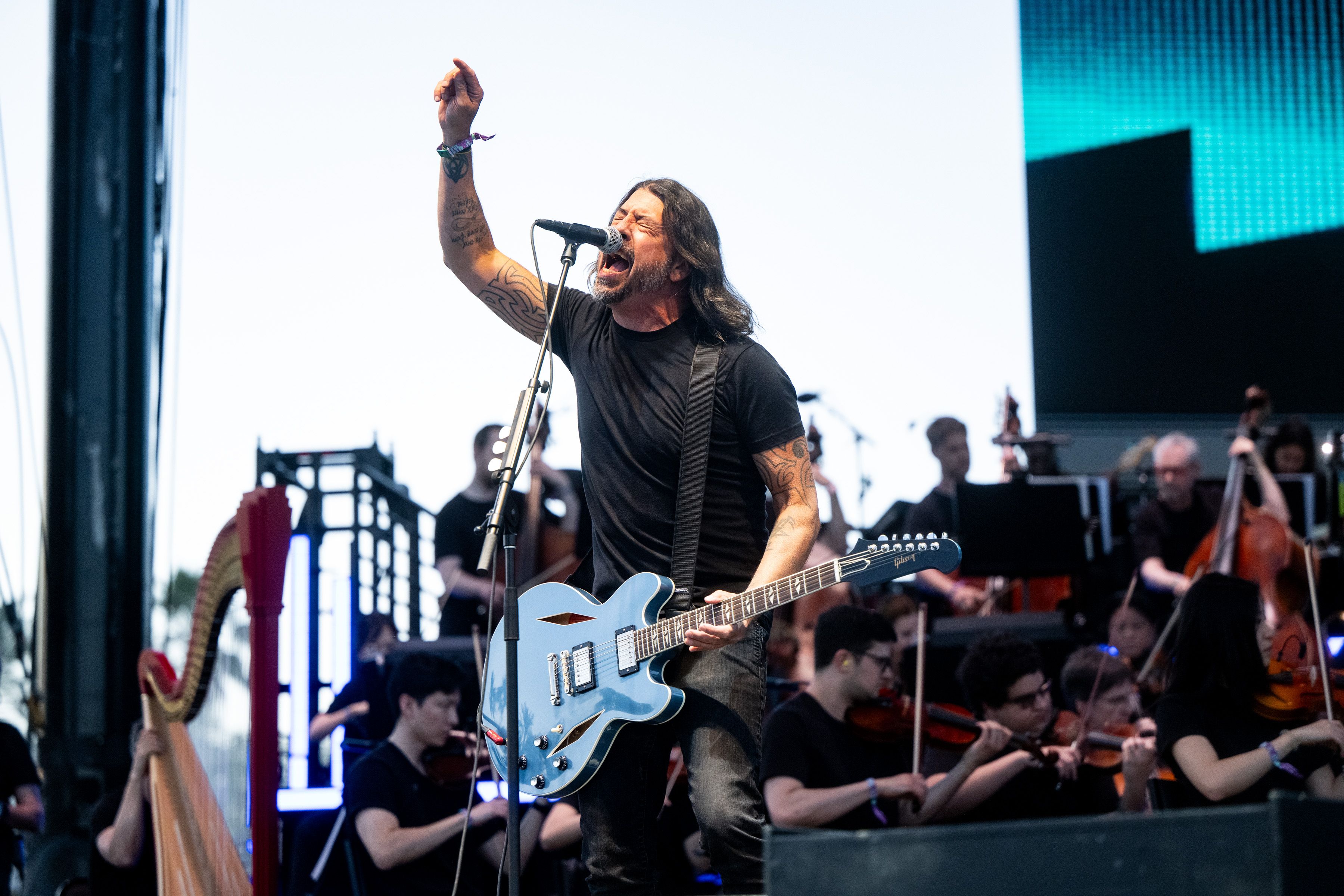 Long-haired musician in black shirt passionately singing and playing light blue electric guitar on stage with orchestra in background during outdoor concert.