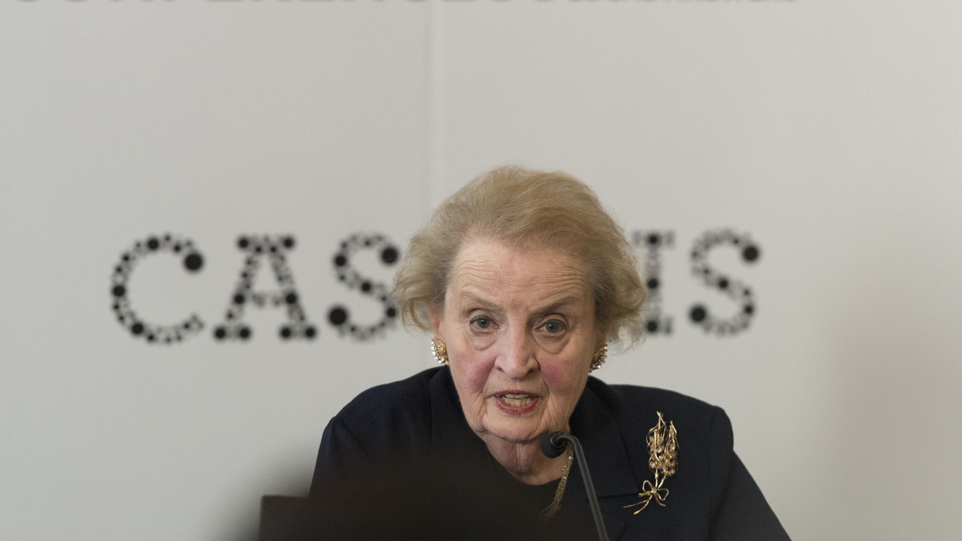 Madeleine Albright in black before a white background with black lettering.