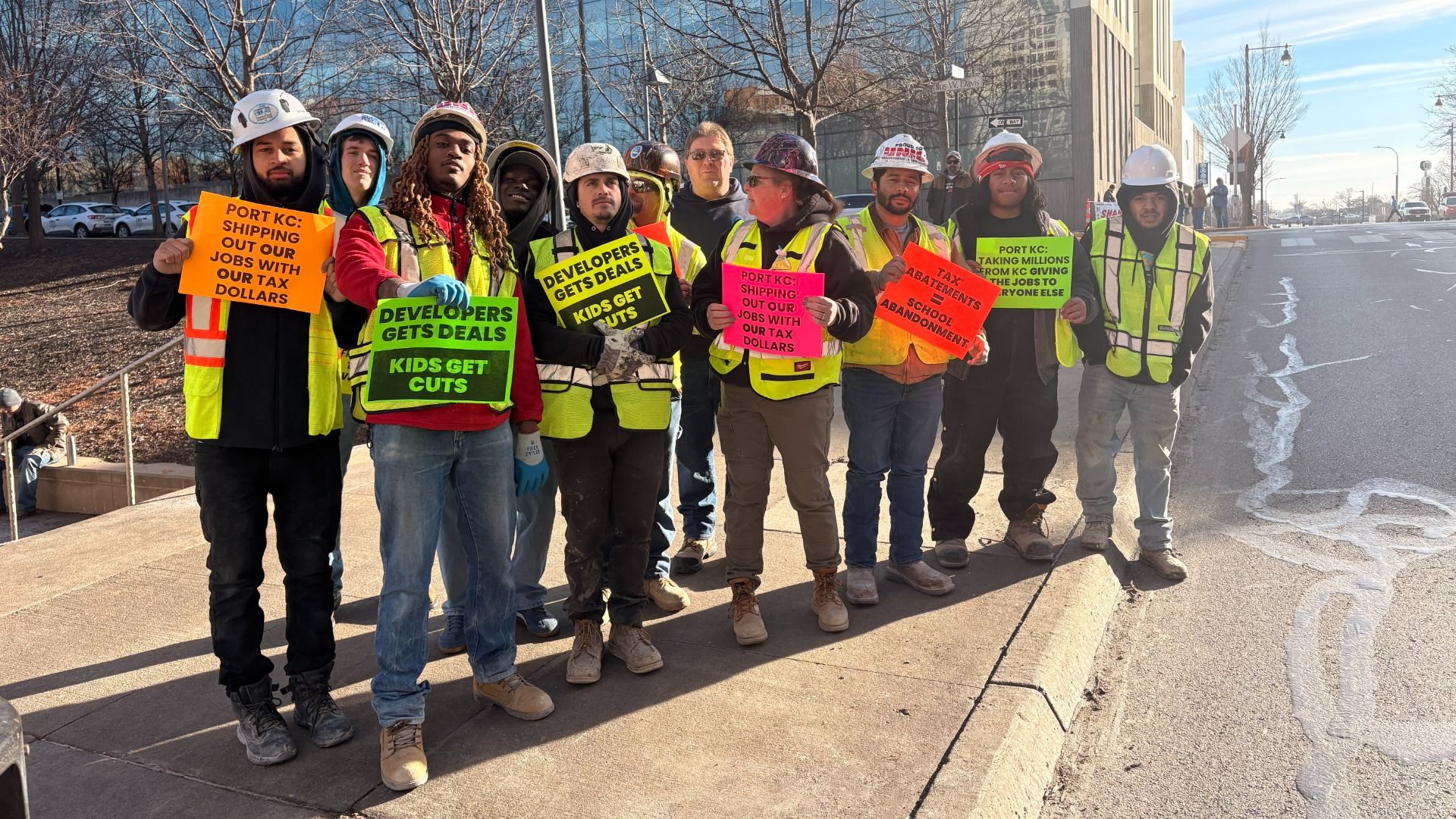 Group of workers in safety vests and helmets holding brightly colored protest signs against tax deals favoring developers over jobs and schools in Port KC, outdoors on a city street.