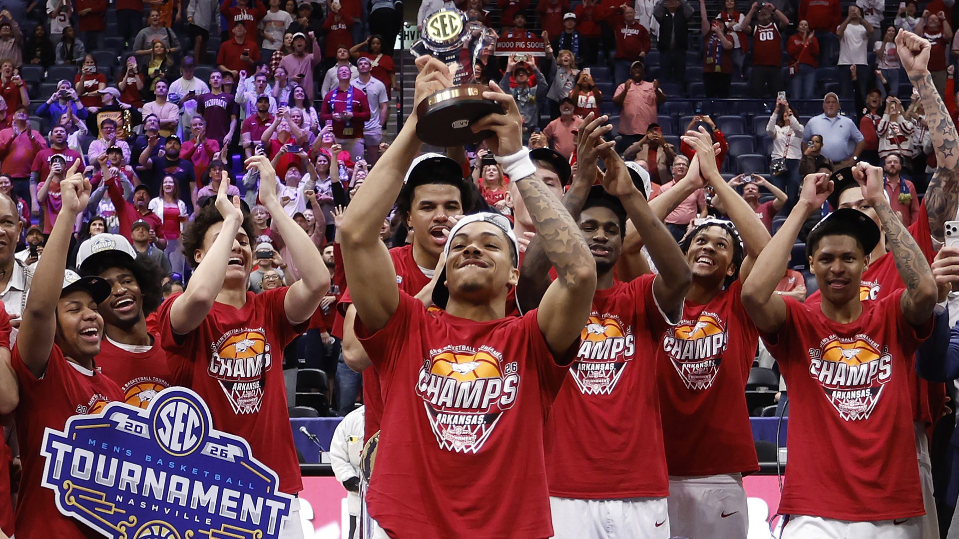 College basketball team in red jerseys lifts a trophy above their heads, celebrating in a crowded arena; fans cheer in the stands, and a trophy sits on the table in the foreground.