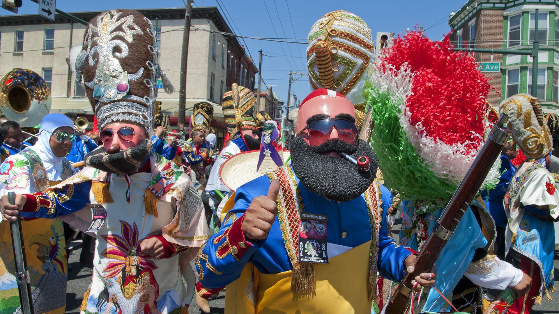 Colorful folkloric parade with people wearing elaborate embroidered costumes, tall decorated hats, red-tinted sunglasses, fake beards, and carrying props, set on a city street under blue sky.