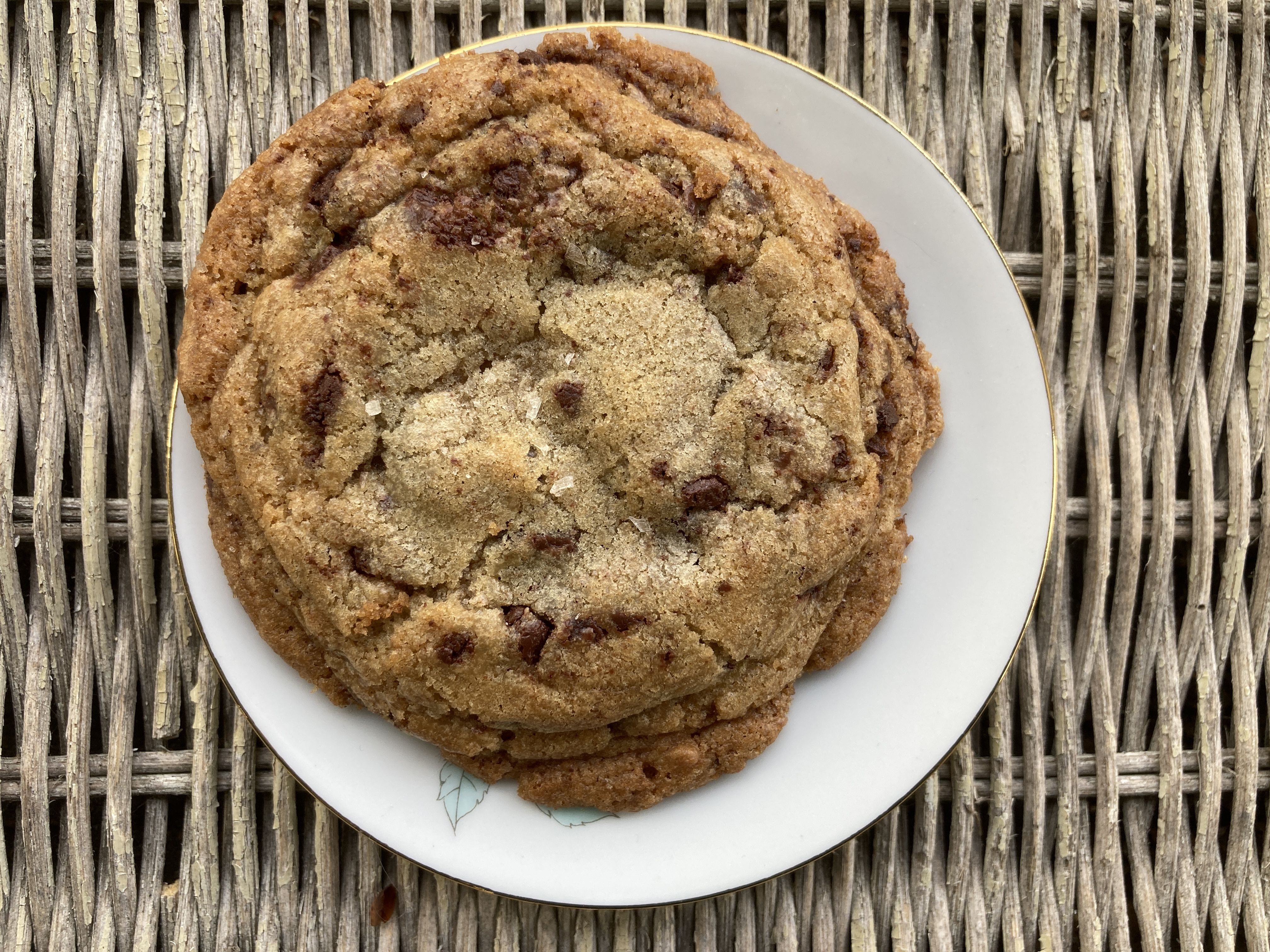 Taken from above; the top of a thick and nubbly chocolate chip cookie, on a small white plate on a wicker table.
