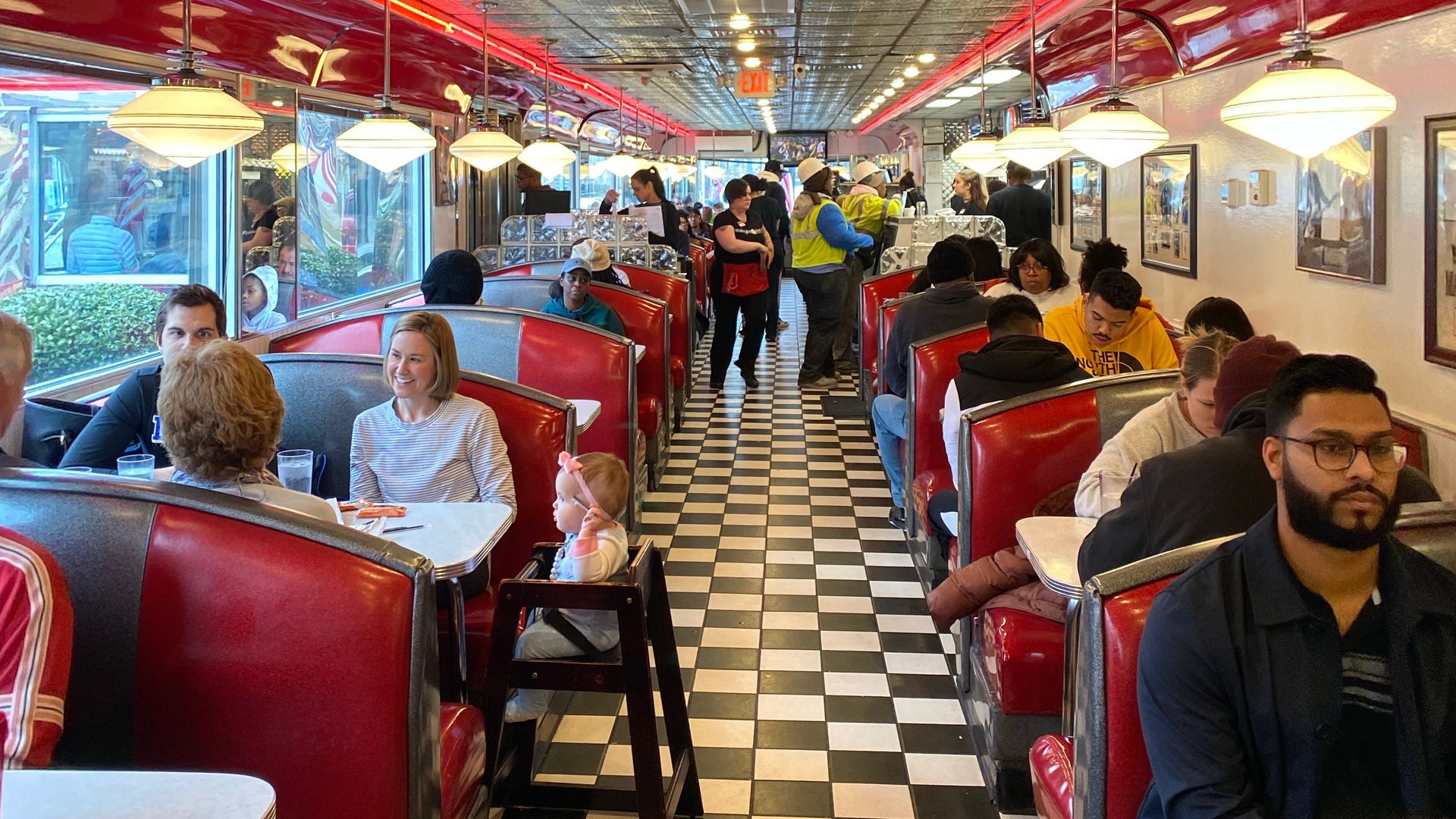 Busy retro diner interior with black-and-white checkered floor, red booths, silver trim, pendant lights, and customers seated or standing along the aisle.