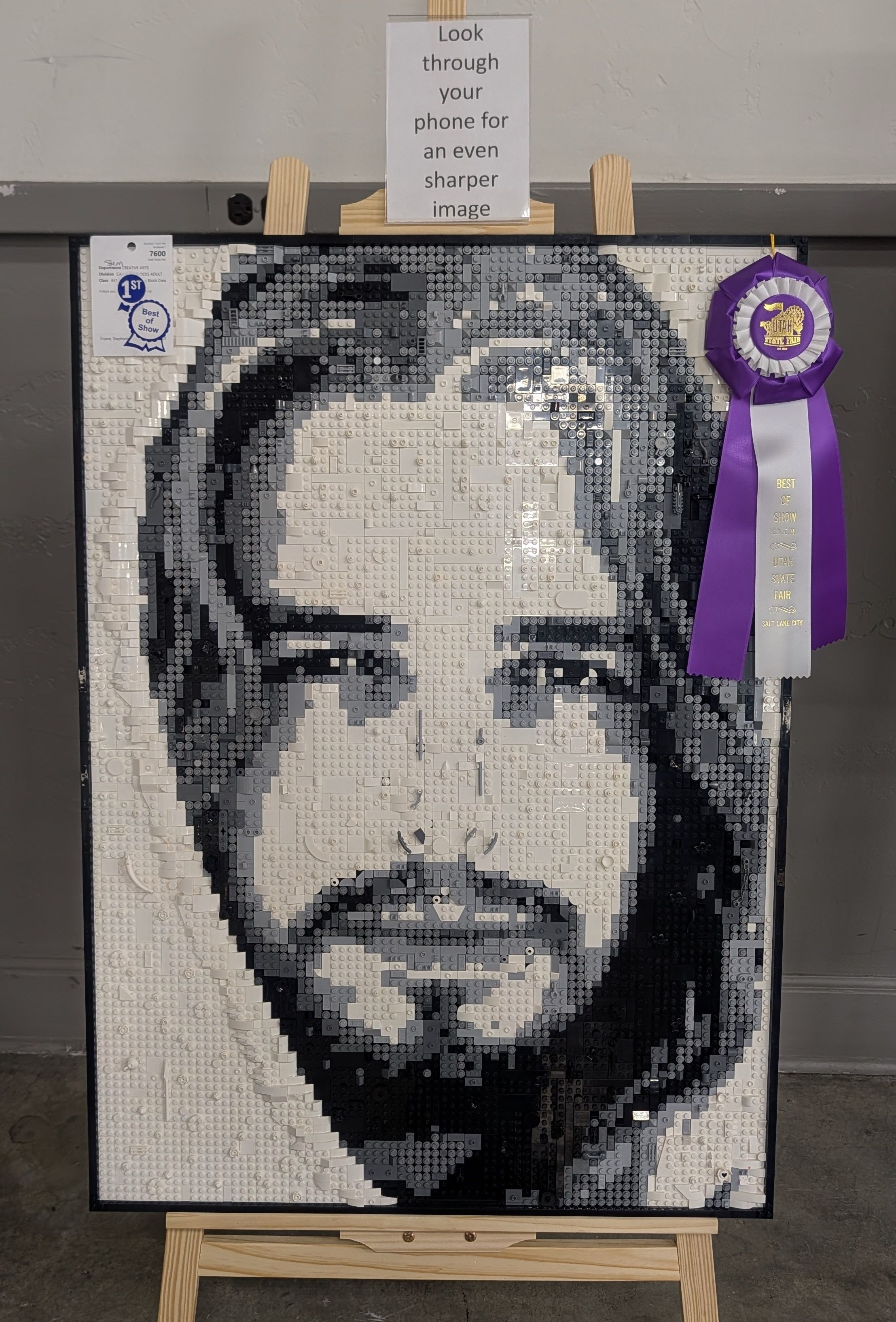 Large grayscale LEGO mosaic portrait on a black frame, displayed on an easel. A sign above reads, "Look through your phone for an even sharper image," with a purple Best of Show ribbon hanging to the right.