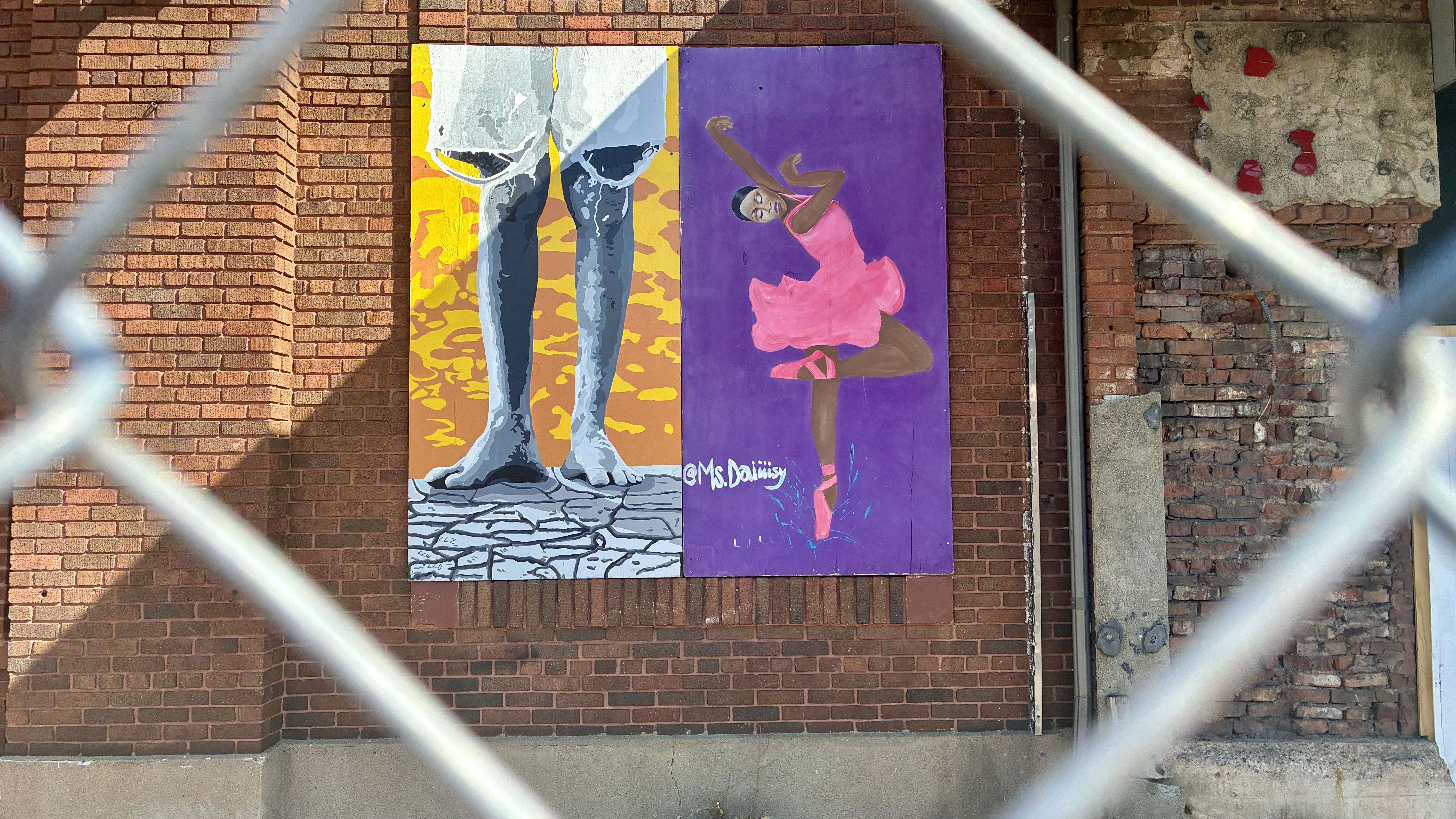 Seen through a chain-link fence, a two-panel mural on a brick wall: left shows jeans on cracked ground with a yellow-orange backdrop; right shows a dancer in a pink dress against purple, signed