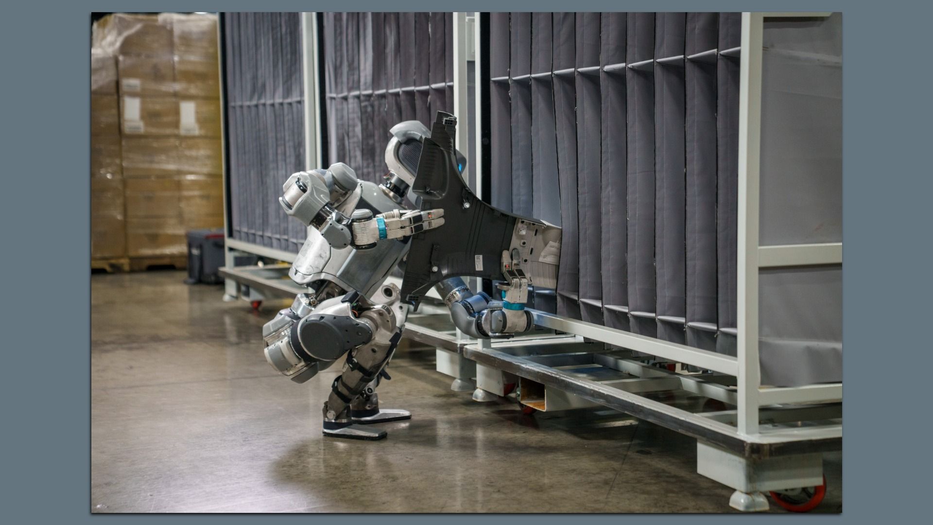 A silver humanoid robot squats while inserting a large black vehicle part into a metal rack in an industrial setting with a concrete floor.