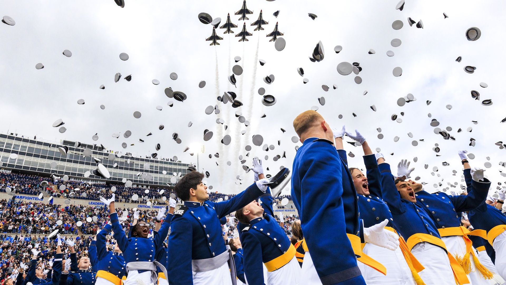 Graduates in blue uniforms and white gloves celebrate by throwing gray mortarboards into a cloudy sky over a packed stadium, as jets leave white smoke trails behind them.