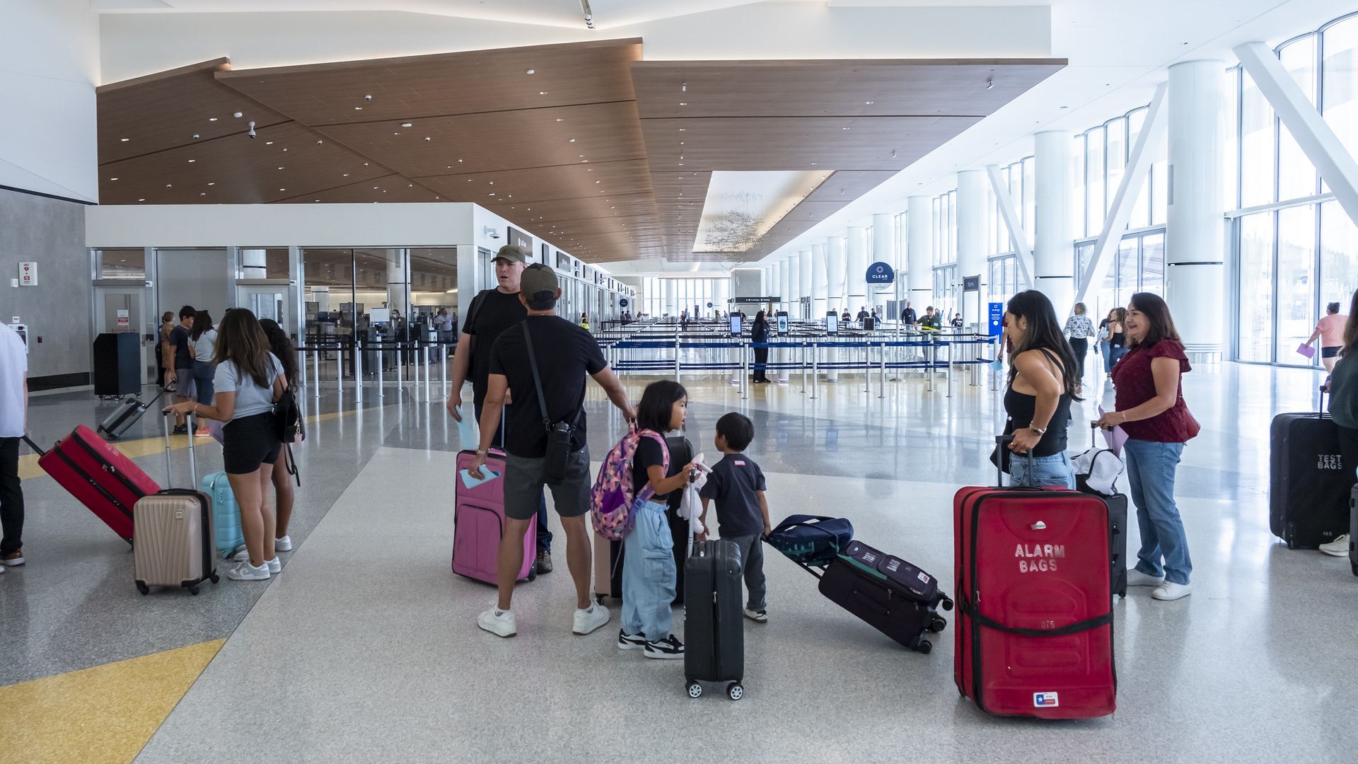 People with colorful suitcases, including pink and red, wait in a bright, modern airport terminal with high ceilings and large windows near security checkpoints.