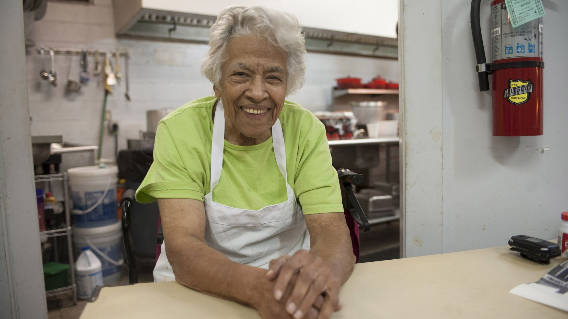 Photo shows chef Leah Chase sitting and smiling at a table by the kitchen in her restaurant. She's wearing a green t-shirt and a white apron. 