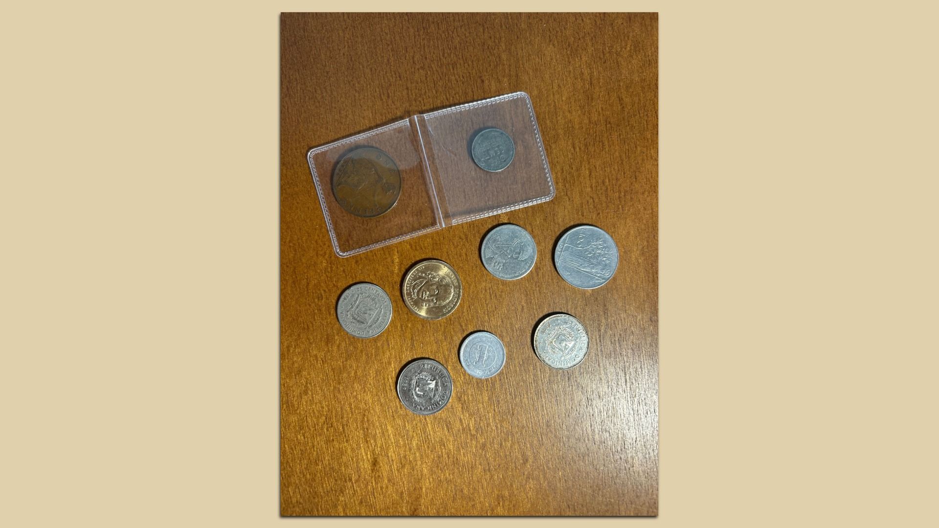 Various coins of different sizes and colors, including silver and gold tones, displayed on a wooden surface with two coins in a plastic holder at the top.