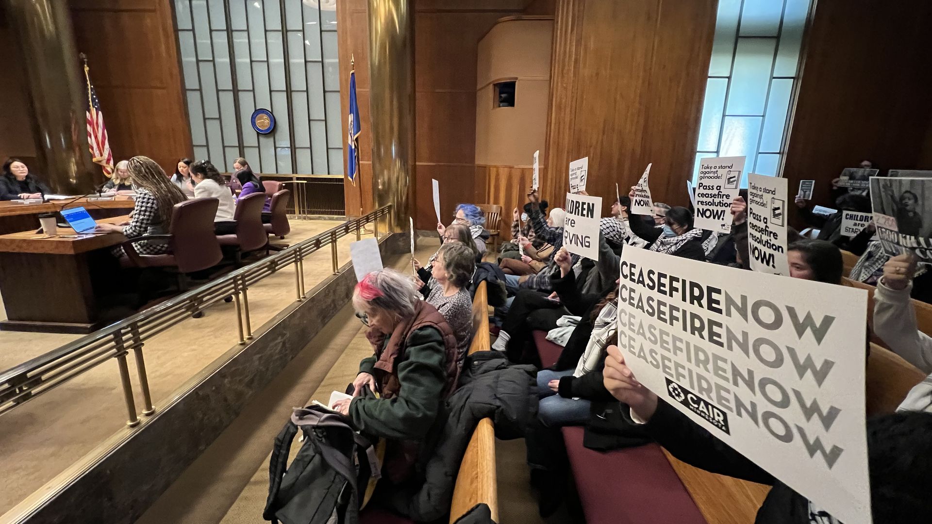 A sign saying "Ceasefire Now" in a government meeting space with wooden paneling and a city council seated in the background.