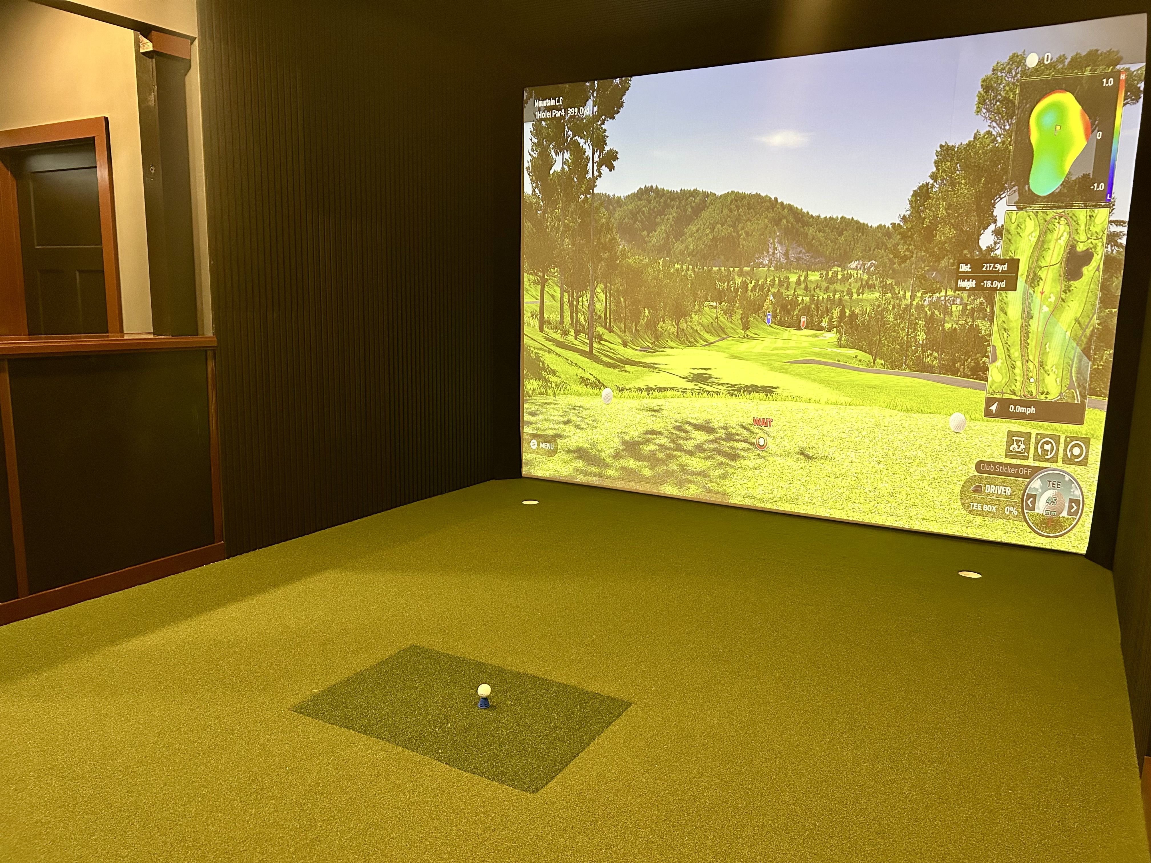 iron oak cigar lounge. Indoor golf simulator: a wide, bright screen shows a lush golf course with trees; a white golf ball on a blue tee rests on a mat in a dim room with dark wood paneling and a door on the left.