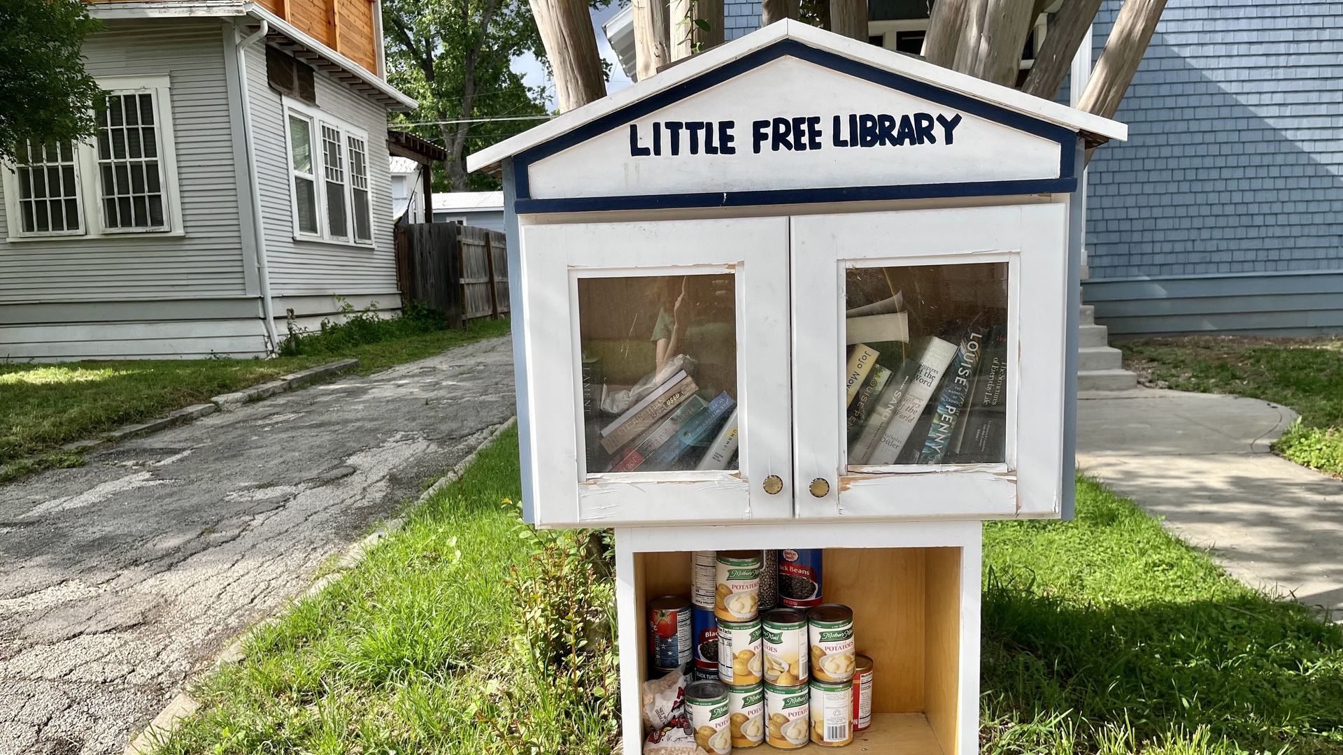 A little free library with a pointed roof, glass doors for the book case and an open area with canned food on the bottom.
