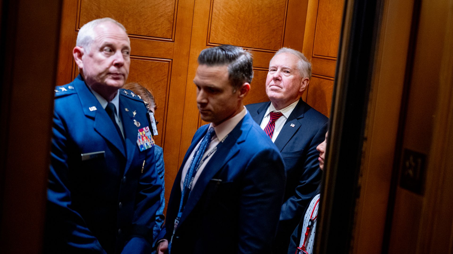 Five people stand in an elevator. The tone is warm. Three of the people, men, are in blue clothing, including one in a military uniform.