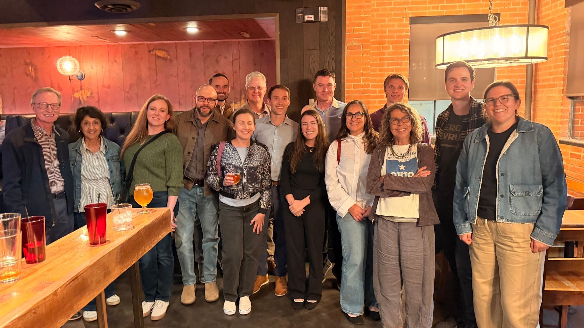 Group photo of about 15 adults in a brick-walled bar, smiling at the camera. Drinks sit on a wooden bar in the foreground; warm lighting and casual attire.