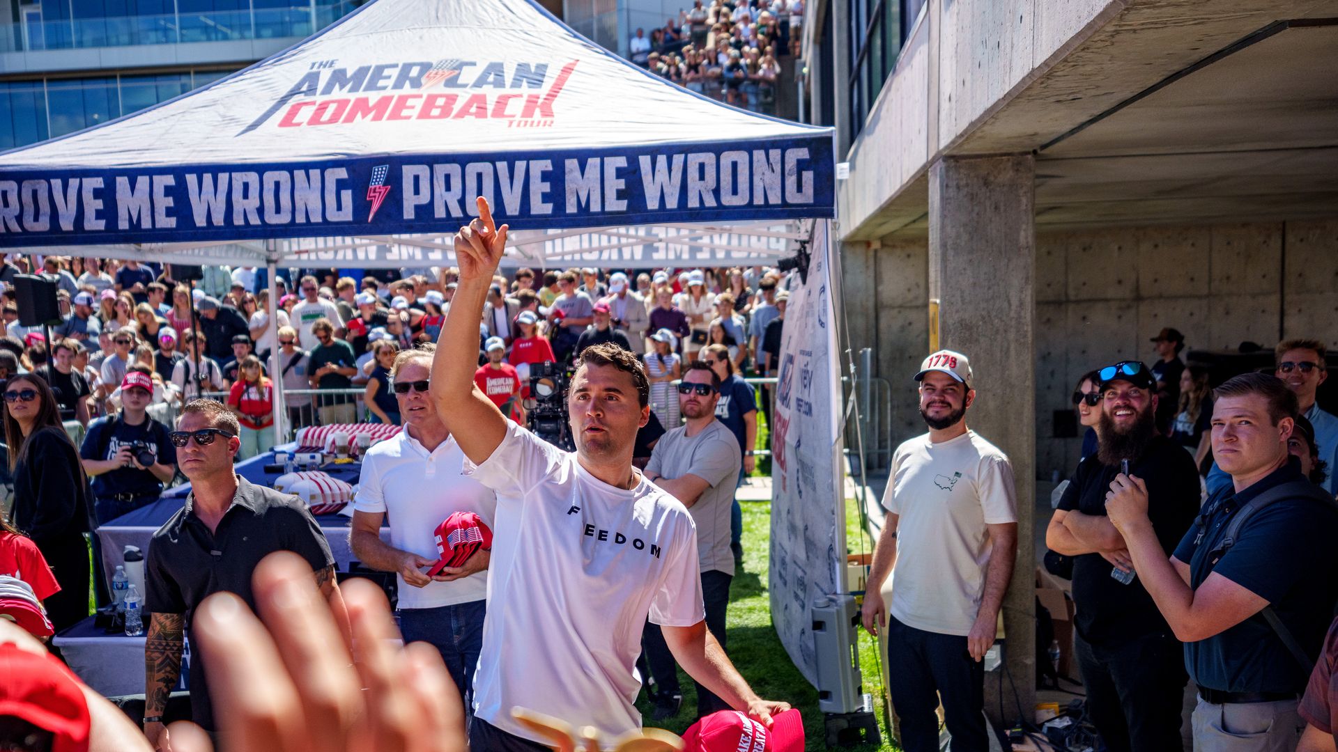 Charlie Kirk holds red MAGA hats in one hand while throwing them to a crowd with the other. Behind him, a tent reads "American Comeback" and "prove me wrong." 