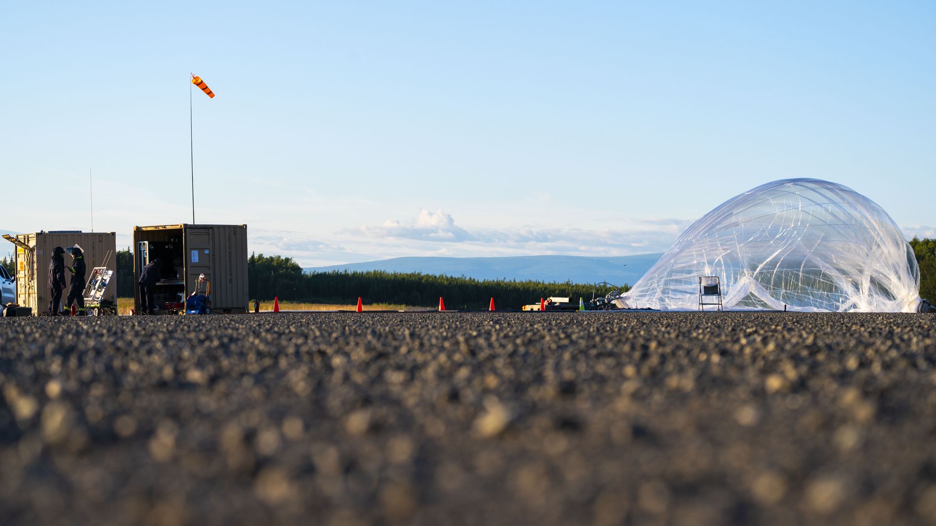 Low-angle view of an outdoor setup with a transparent large balloon partially inflated, two small brown containers, orange cones, and a windsock against a clear blue sky and forest background.